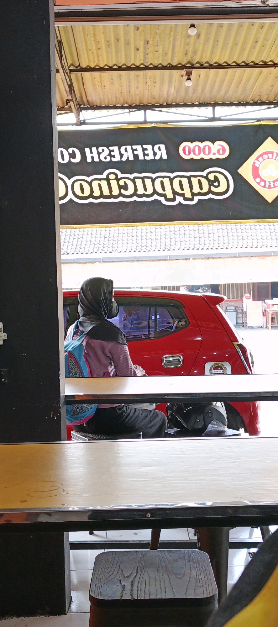 In Sidoarum, Indonesia, a person wearing a headscarf and mask is seated in a red car. They are looking away from the camera. The car's interior is not visible. A sign in the background displays "CAPPUCCINO COFFEE" and "000.e" in large white letters against a black background. The sign is partially obscured by a corrugated metal roof overhead. The foreground shows a wooden table and a stool.