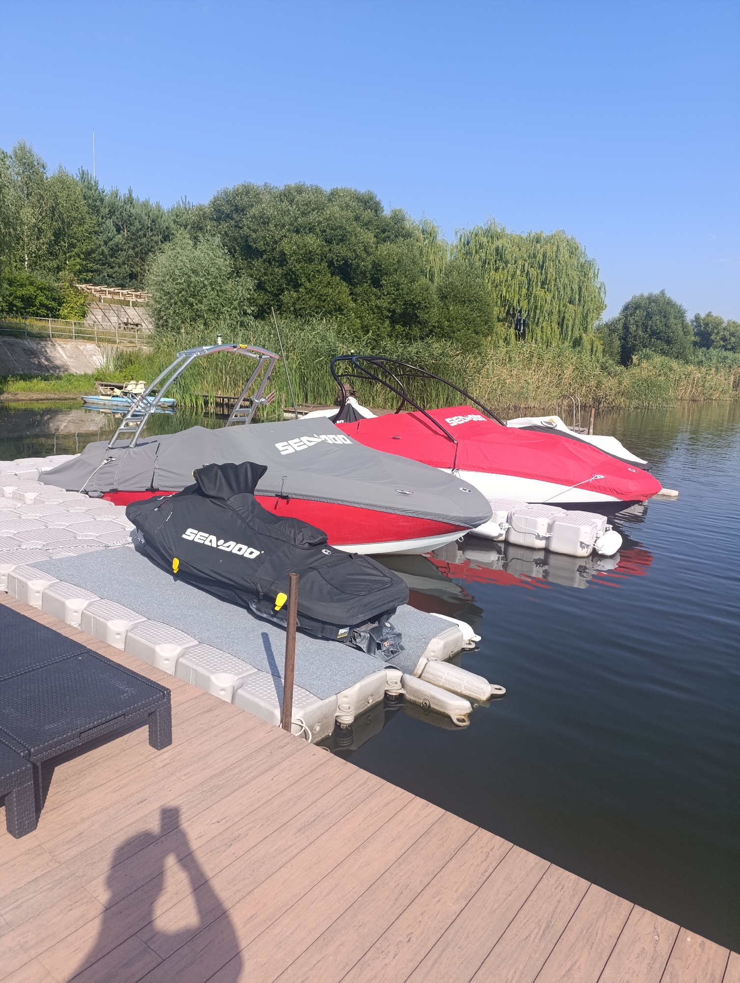 In Bulatselivka, Ukraine, a serene lakeside scene unfolds under a clear blue sky. Three personal watercraft are docked on a modular floating platform. The closest watercraft is covered with a black cover displaying the "SEA-DOO" logo in white. Adjacent to it, another watercraft is partially visible, covered in a grey tarp. A third, red watercraft with a white hull is moored further back, also under a red cover. The platform is made of interconnected grey and white plastic modules. A black mesh lounge chair sits on the wooden deck in the foreground, to the left of the frame. The water is calm and reflects the blue sky and surrounding greenery. In the background, lush trees and reeds line the shore, with a distant structure visible on the left bank. A shadow of a person holding a phone is cast on the wooden deck, indicating the photographer's presence.
