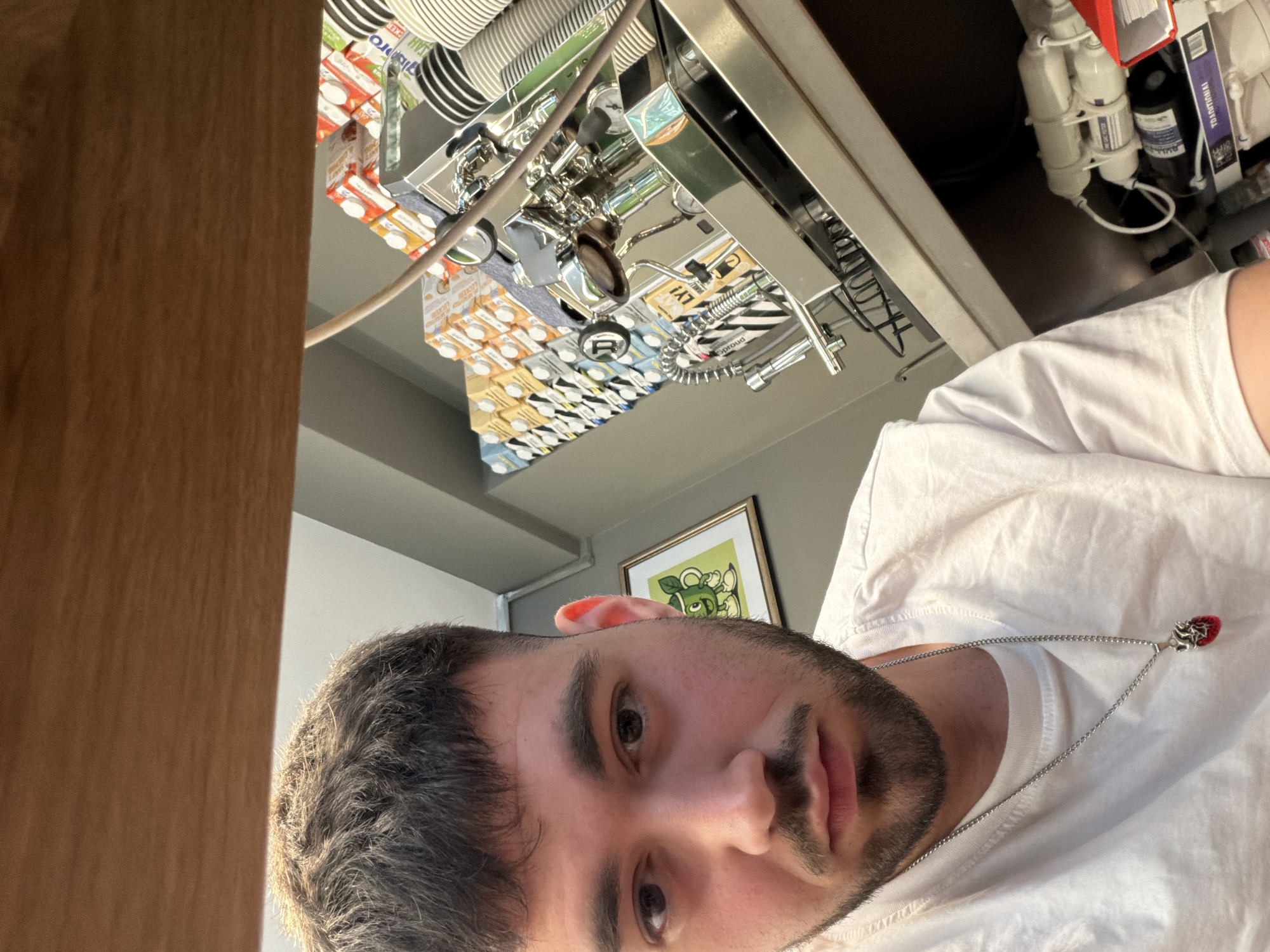 An indoor, eye-level, close-up selfie taken by a light-skinned male with dark hair, a beard, and a white t-shirt. He is wearing a silver chain with a red heart-shaped pendant. Behind him, to the right, is a stainless steel espresso machine. Stacked behind the espresso machine are numerous small cartons of what appear to be milk or juice. To the left of the espresso machine is a framed picture on the wall of a cartoon green character. The environment appears to be a cafe or coffee shop. The specific location in Sofia, Bulgaria, cannot be determined from the image.