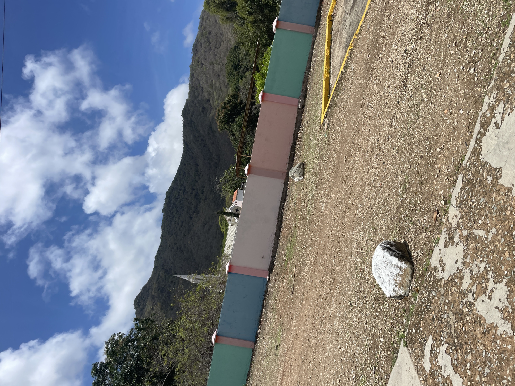 The image captures a scenic view under a bright, partly cloudy blue sky. In the foreground, a gravelly ground surface is visible, interspersed with patches of concrete and sparse green vegetation. A large, light-colored rock rests on the gravelly area towards the left of the frame. A yellow-lined concrete barrier or parking divider is also present in the lower right corner.
A multi-colored wall stretches horizontally across the mid-ground, divided into sections of light green, blue, pink, and white, with pink decorative pillars. Beyond the wall, several buildings with white roofs are visible, including a prominent white building with a tall, slender steeple, suggesting a church or religious institution. A dense, green mountainous landscape forms the backdrop, with trees dotting its slopes. The overall scene depicts an outdoor environment, likely in a town or village, with a clear emphasis on the natural landscape and man-made structures.
This location is identified as being in El Valle del Espíritu Santo, Venezuela.