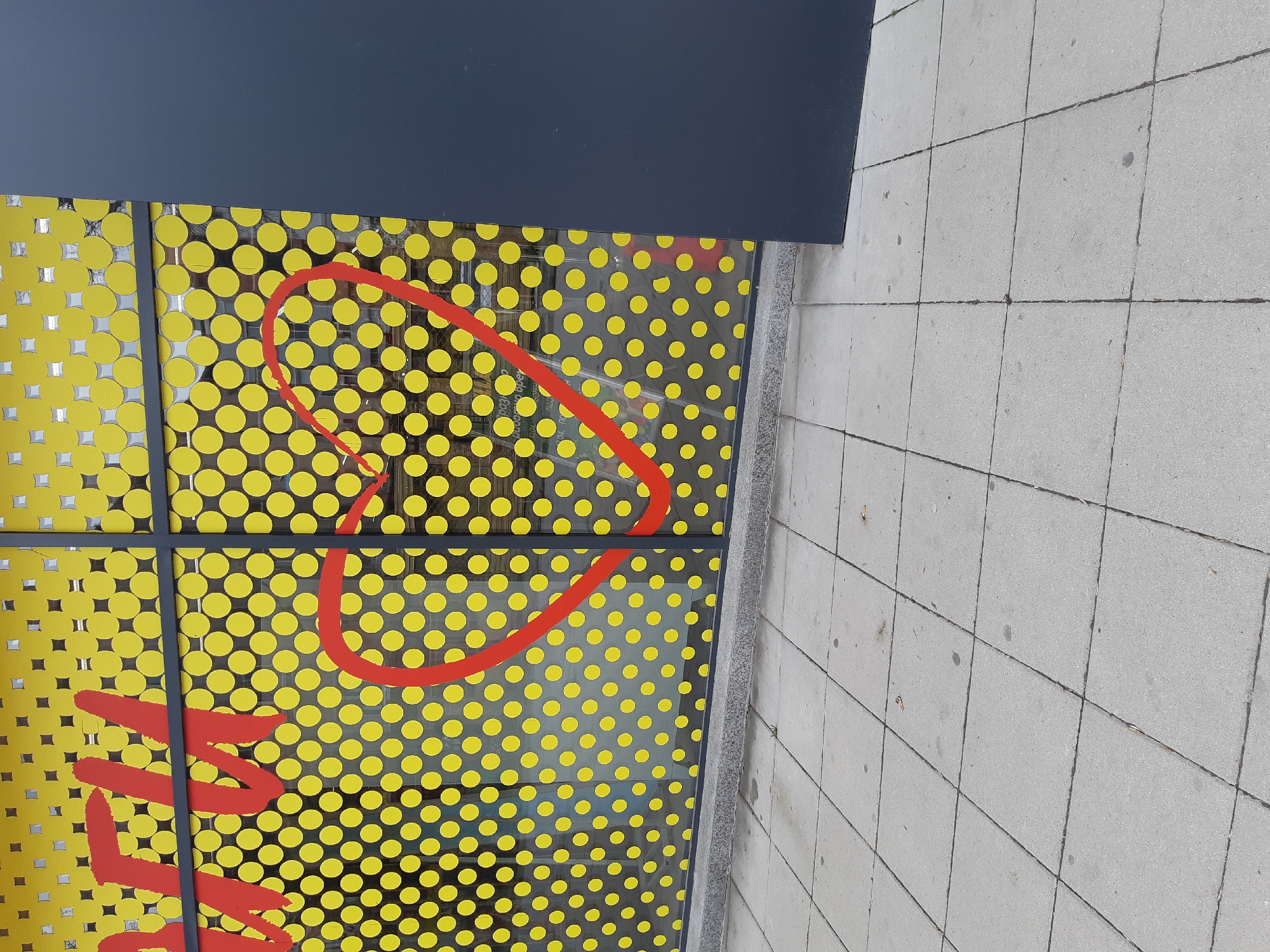 A ground-level, eye-level shot depicts an outdoor scene featuring the facade of a building, likely a store or establishment, in Plovdiv, Bulgaria. The facade is covered in a yellow background overlaid with uniformly spaced yellow circles and overlaid in red, a heart shape and letters. The building facade is adjacent to a concrete pavement. The angle of the image suggests it was taken from ground level. The photograph is taken outdoors.