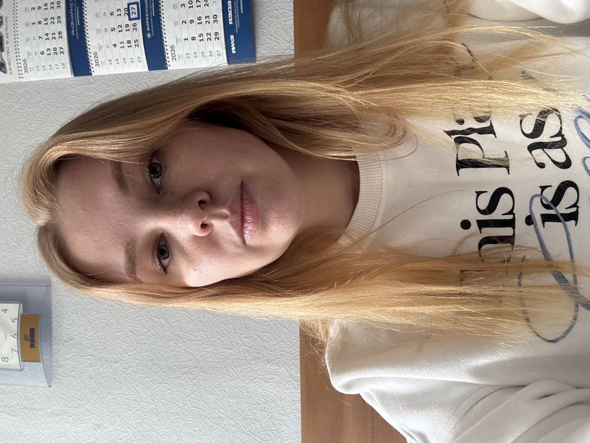 The image depicts a young woman with long, blonde hair, wearing a white sweatshirt with black and blue lettering. Her face is in focus, revealing fair skin, grey eyes, and subtle makeup. To the left of the frame, a calendar is visible, showing dates in 2026, with Russian text. A flip-style desk calendar displays the date as the 5th. The background consists of a textured, light-colored surface, possibly a wall, and a wooden surface.
It is impossible to verify the location in the city of Rusanovo, Russia using only this image.