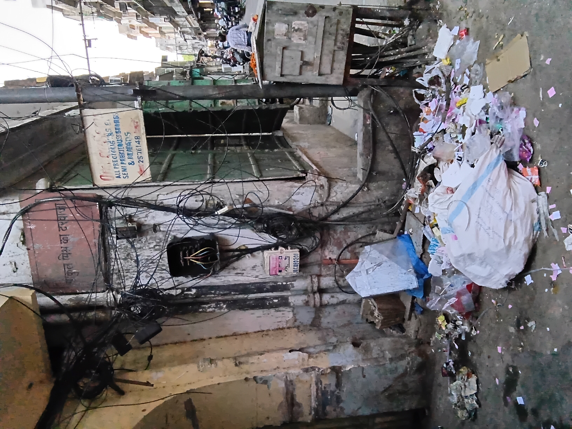 The image depicts a narrow alleyway in Jaipur, India, characterized by overhanging electrical wires, exposed electrical boxes, and accumulated trash. The alley is flanked by weathered buildings with peeling paint. An advertisement for "ALL PRECIOUS & SEMI PRECIOUS STONES & BEADS" with the phone number 2576148 is visible. Another sign reads "कुल मिश्र का दरवाजा" and "JKC080201". A person on a motorcycle is visible in the background. The alleyway is littered with plastic bags and assorted debris.