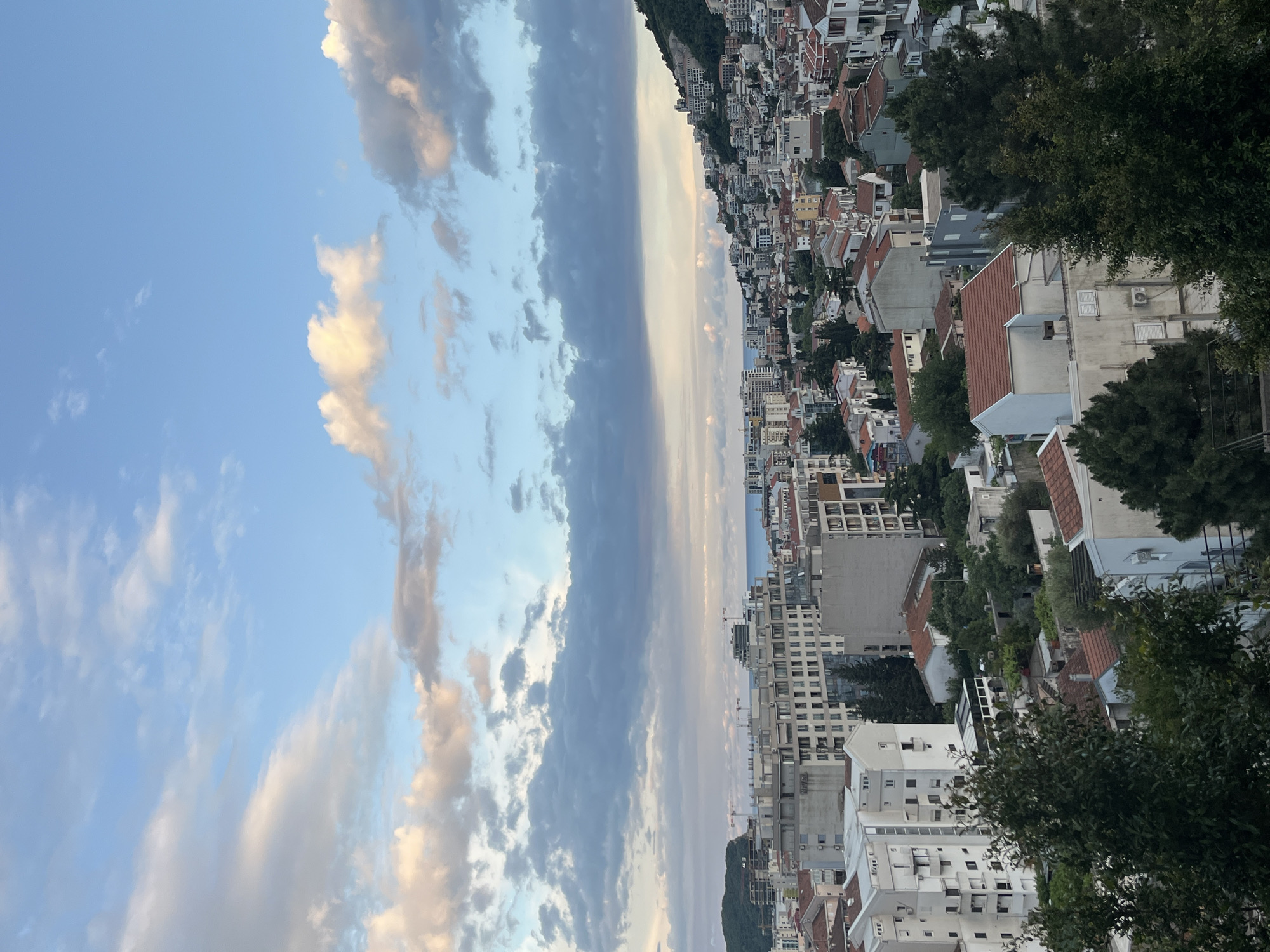 A slightly rotated eye-level long shot captures a cityscape under a sky with a blend of blue and gray clouds. The city, which is Budva, Montenegro, features buildings of varying heights and designs, predominantly white and beige with red-tiled roofs. The structures are densely packed, extending toward the horizon where a sliver of water is visible. The lower part of the frame shows the tops of trees, adding a touch of green to the urban landscape. The sky is a mix of clear blue and clouds with a mix of white and gray. Some clouds have light edges, suggesting the presence of the sun, possibly near sunset or sunrise.