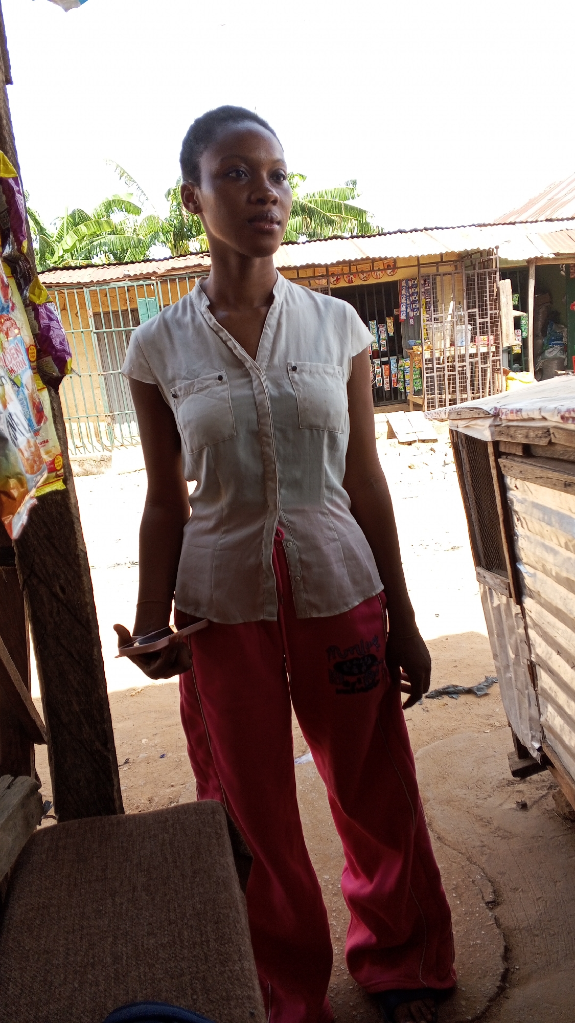 A young woman stands in front of a small, open-air shop in Sabon Tasha, Nigeria. She wears a light-colored, short-sleeved shirt and pink sweatpants with writing on the left leg. The shop behind her has a corrugated metal roof and a display of goods behind bars. A wooden structure with packaged snacks hangs to the left of the woman. The ground is dirt. She is holding a pink phone.