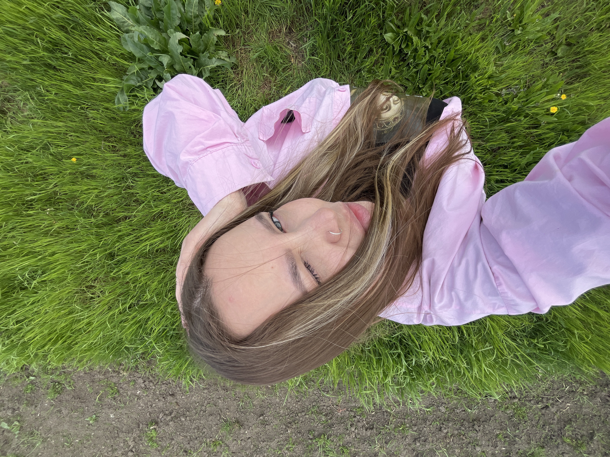 A young woman is depicted lying on her back in lush green grass, viewed from directly above. She is wearing a light pink long-sleeved shirt or jacket, which contrasts with the vibrant green surroundings. Her head is angled slightly, with her light brown hair, featuring lighter highlights, fanned out around her. Her face is visible, showing light blue eyes that are open and looking slightly upwards, away from the camera. She has a small silver nose ring on her left nostril. Her expression appears relaxed and calm, with her lips slightly parted.
Next to her head and upper body, a book or similar object is partially visible. It features an artistic depiction of a face with ornate, gold-toned headwear or jewelry. No legible text is visible on this object.
The setting is outdoors in what appears to be a field or grassy area in Novopokrovskaya, Russia. The ground is predominantly covered in dense, bright green grass. Scattered within the grass are a few small yellow dandelion flowers. Towards the edges of the frame, particularly at the bottom and left, patches of dark, bare soil are visible. There are also larger, darker green plants, possibly dandelions or other wild foliage, in the upper portion of the image. The lighting is soft and diffused, suggesting a daytime scene with either an overcast sky or ambient light, contributing to a peaceful atmosphere. The overall activity is one of repose or quiet contemplation in nature.