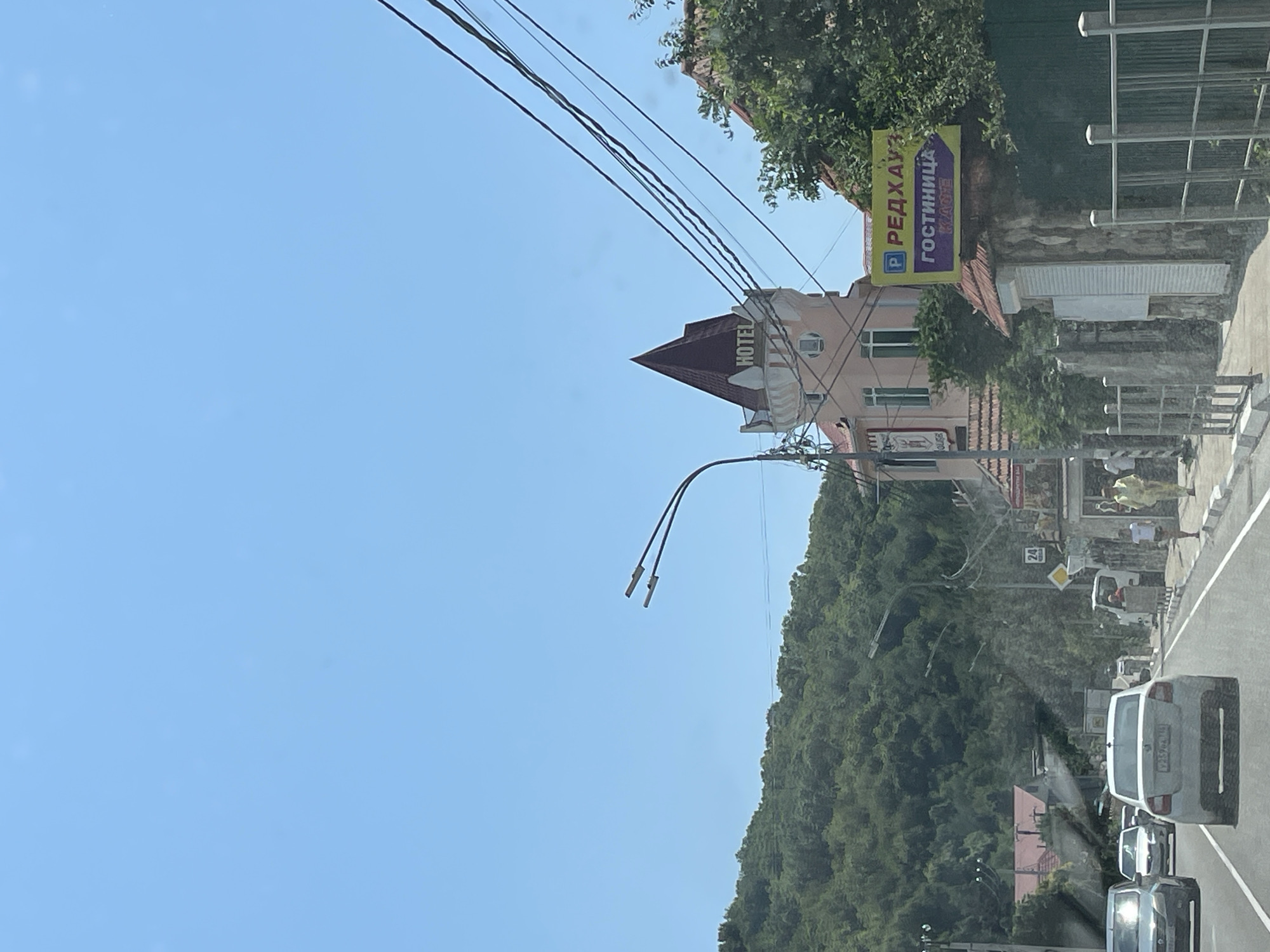 The image depicts a street scene in Shepsi, Russia, on a clear, sunny day. In the foreground, a paved road runs towards the viewer, with several cars driving or parked along it. The dominant structure in the middle ground is a multi-story building with a pink facade and a distinctive red conical roof, featuring the word "HOTEL" prominently displayed. To the right of the hotel, a yellow sign with blue and purple lettering indicates "РЕДХАУЗ ГОСТИНИЦА," suggesting a hotel named "Redhouse." A lush green, forested hill forms the backdrop of the scene. Power lines are visible strung across the sky. Several pedestrians are present on the sidewalk, some walking and others standing. The architecture suggests a resort town, likely along the Black Sea coast, given Shepsi's location.