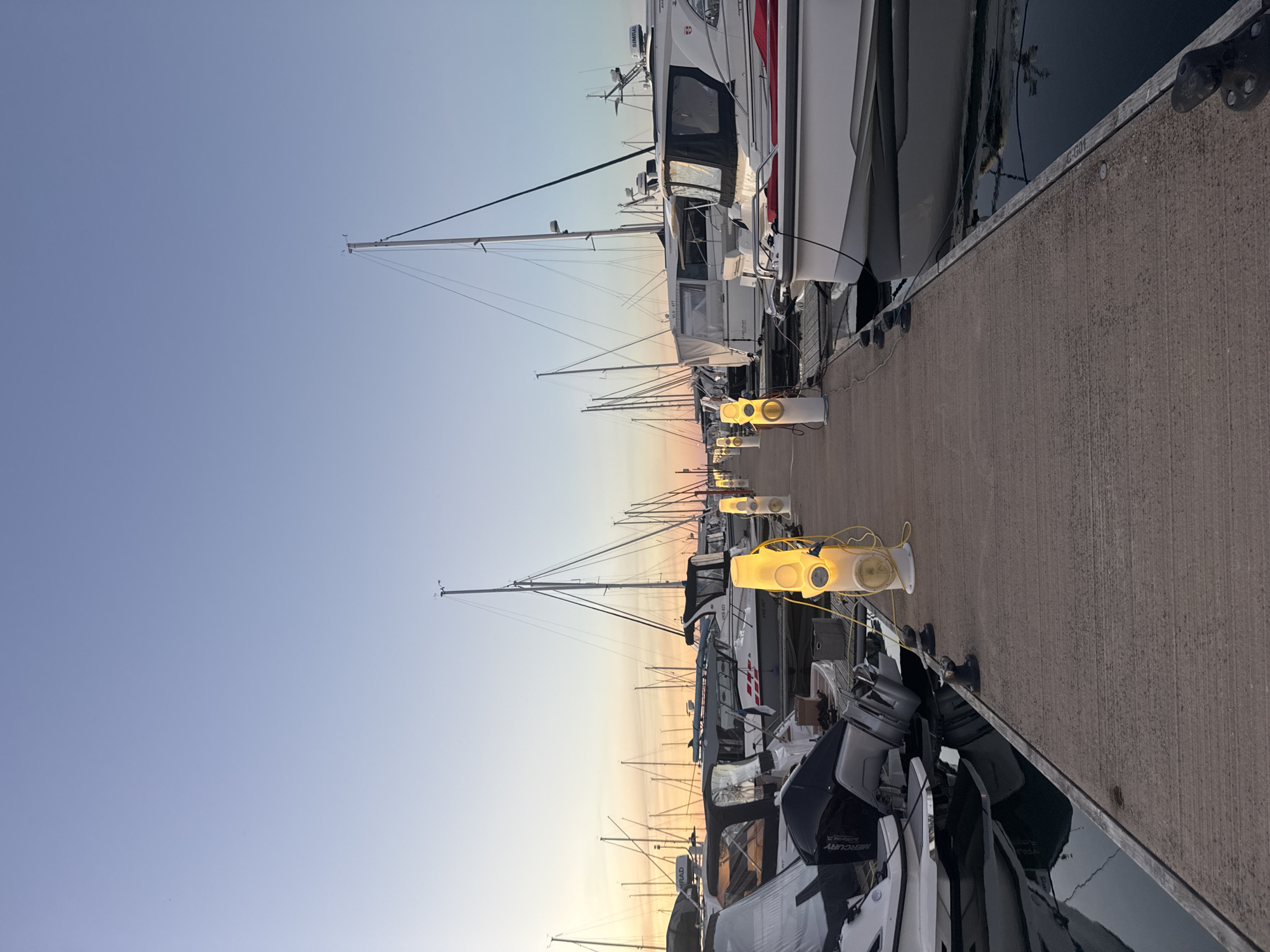 The image depicts a marina in Tallinn, Estonia, during twilight. A concrete pier runs through the center of the frame, lined with docked boats on both sides. The boats are of various sizes, including sailboats and motorboats, some covered with tarps. Several modern, yellow bollard-like lights are spaced along the pier, illuminating the pathway. The sky above is a clear gradient of pale blue and soft orange, indicative of a sunset or sunrise. The water in the marina is dark and calm, reflecting the sky and the lights. There are no people visible in the image.