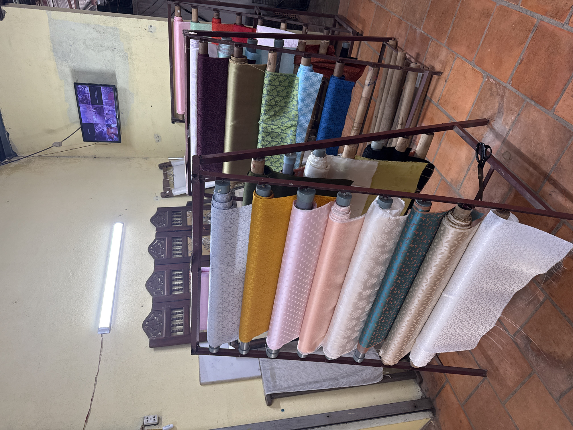 The image displays an indoor commercial space, likely a fabric shop or workshop, in Nam Ban, Vietnam. The central focus is on numerous rolls of textiles neatly arranged on multi-tiered metal racks. The fabrics exhibit a wide array of colors and patterns, including vibrant pinks, purples, golds, greens, blues, teals, whites, and creams. Many rolls feature intricate woven patterns, suggesting silk or brocade fabrics.
The floor is covered with reddish-brown terracotta tiles, while the walls are painted a pale yellow or cream color. On one wall, a flat-screen monitor is mounted, showing a grid of surveillance camera feeds, indicating security monitoring within the establishment. An electrical outlet with visible plugs is situated near the bottom of this wall. A long, vertical fluorescent light fixture illuminates the space from another wall, with its wires visibly descending.
A decorative, carved wooden panel, possibly part of a window or architectural detail, is visible on the wall behind some of the fabric racks. A pair of large black scissors hangs from one of the lower metal display bars, implying that fabric is cut on-site. Behind the fabric displays, several wooden poles or dowels are stacked. No people are visible in the scene. The lighting suggests it could be any time of day indoors, under artificial illumination. The overall setting appears functional and dedicated to the sale or creation of textiles.