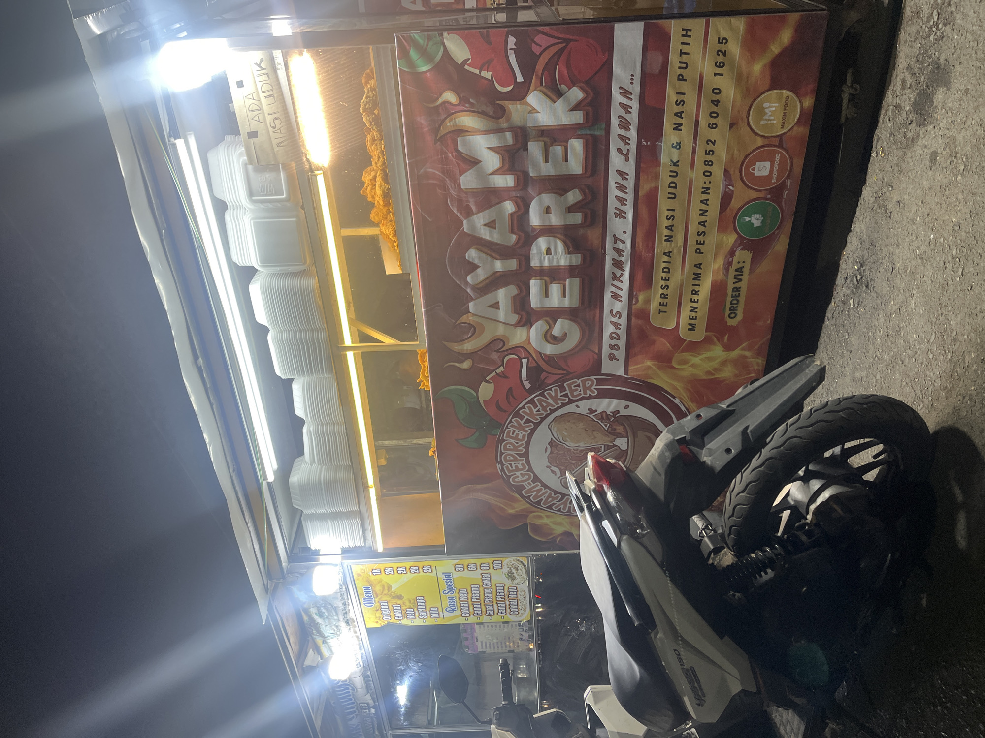 The image shows a food stall selling "Ayam Geprek" and "Nasi Uduk" in Banda Aceh, Indonesia, at night. The stall has a brightly lit counter displaying stacks of white takeout containers and a sign with the text "AYAM GEPREK". Below this, another sign reads "PEDAS NIKMAT, HANA LAWAN..." (Spicy, Delicious, No match...). Further down, it states "TERSEDIA NASI UDUK & NASI PUTIH" (Nasi Uduk & White Rice Available) and provides a phone number for orders: "0852 6040 1625". There are also logos for food delivery services: GrabFood, ShopeeFood, and Maxim Food. A scooter is parked in front of the stall. The lighting suggests it is an outdoor setting, possibly on a street or a local market.