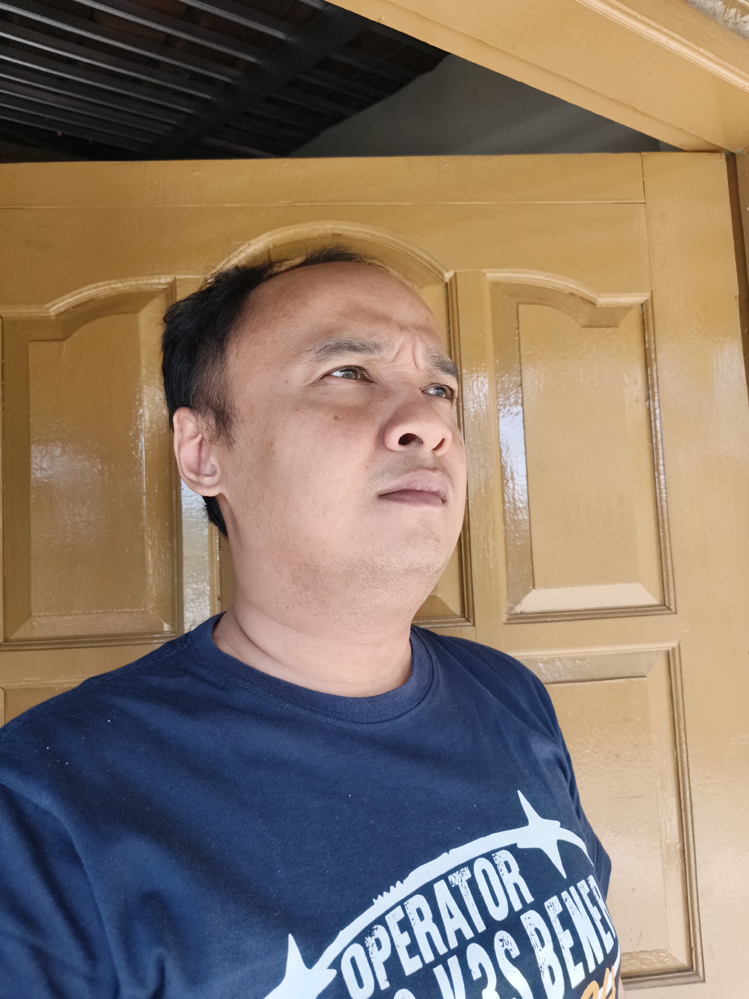 A medium shot captures a man from the chest up, positioned in front of a wooden door with raised panels. The man is looking upwards and to his right, his brow furrowed. He has dark hair, short on the sides and longer on top, and fair skin with some stubble. He is wearing a navy blue t-shirt with white lettering that appears to read "OPERATOR VS BENEFIT" with a graphic of a bird. The door behind him is painted a glossy golden-brown. Above the door, a dark ceiling with exposed metal beams is visible. The overall lighting suggests an outdoor or semi-covered environment. The location is identified as Kaliwader, Indonesia.