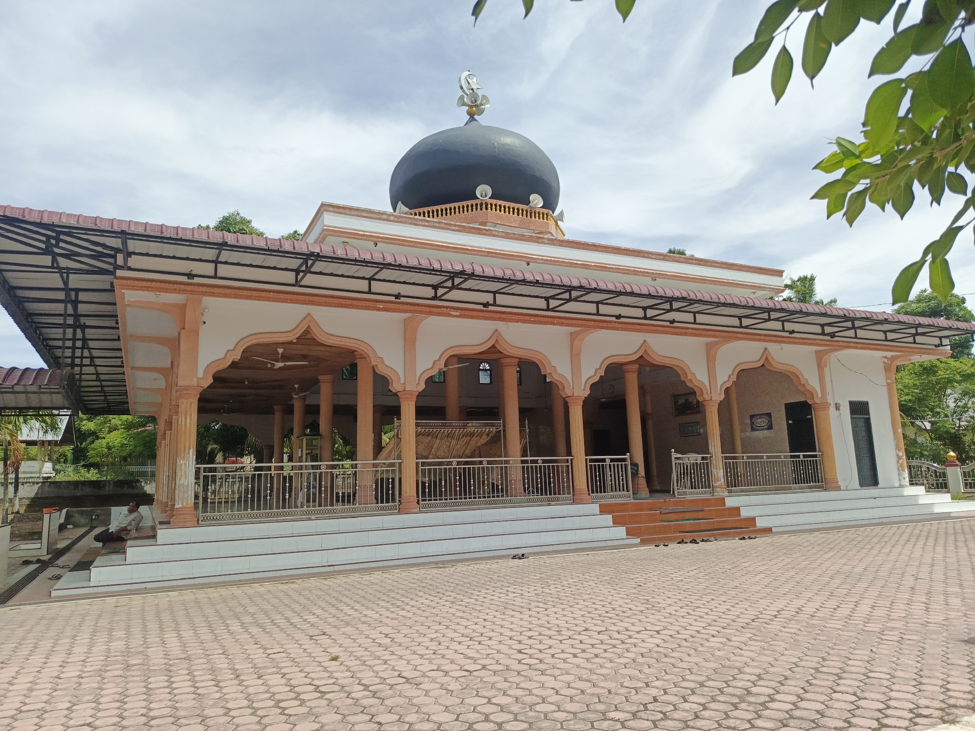 The image shows a mosque in Sawang, Indonesia, with a prominent black dome topped by a crescent moon symbol and speakers. The mosque features an open-air covered veranda with orange columns and white archways, supported by a metal roof structure. A metal railing encloses the veranda. The ground in front of the mosque is paved with hexagonal tiles. To the left of the mosque, a man is sitting on the steps, and several pairs of sandals are neatly placed on the ground. Lush green trees and foliage surround the mosque, and the sky is partly cloudy. The architectural style suggests a tropical climate.