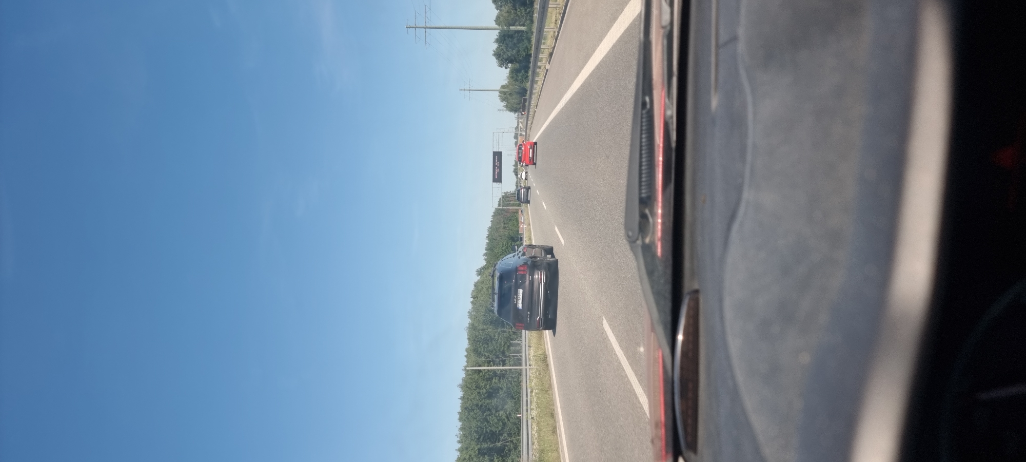 The image shows a view from inside a vehicle looking out onto a highway in Wólka Pracka, Poland. The sky is clear and blue. Several cars are visible on the highway, including a dark SUV in the left lane and a red car in the right lane. The highway is bordered by trees and vegetation on both sides. Overhead signs are present, with one electronic sign displaying text in Polish, likely indicating traffic information or directions. The interior of the vehicle, including the dashboard and windshield, is partially visible in the foreground, suggesting the image was taken while driving.