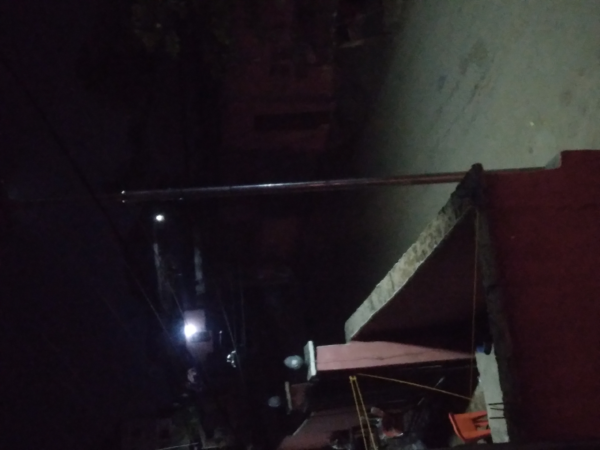 A low-angle nighttime shot appears to be taken in Onitsha, Nigeria. The image's upper right side is dominated by a light-colored concrete or paved area. Below this, there's a brick or concrete structure painted a dark red.
A metal bar or pipe stretches across the center of the frame, seemingly spanning the width of the road or area. Below this, a partial view of buildings or structures, one featuring an angled roof. The lighting is poor and limited, making it hard to identify distinct shapes and features.
The left side of the picture is dark and obscured, with only a streetlight and electrical lines visible in the background. The presence of a streetlight and electrical cables suggests an urban or semi-urban setting. In the very bottom, there's a glimpse of objects, including a partially visible orange chair or container.