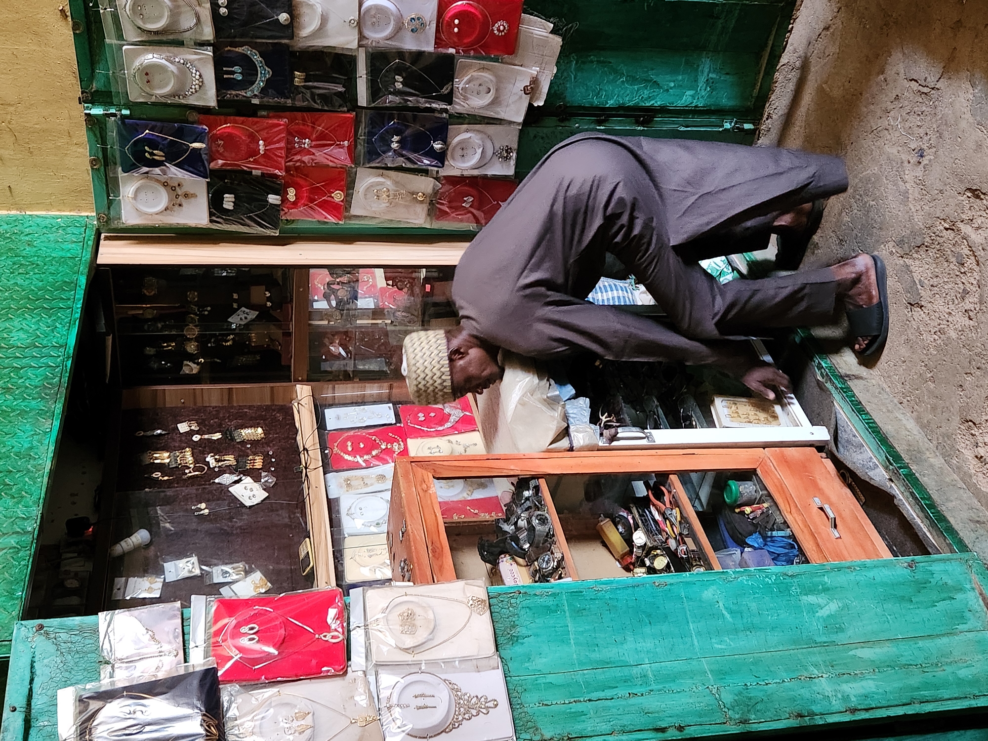 An adult male wearing a light-colored kufi cap and a dark brown kaftan is bent over, facing downward toward a partially opened, green-painted wooden shop display. The shop appears to contain jewelry and watches, including items packaged on white, red, and blue cards. The man's left hand is near a tray of items inside the shop. His feet are clad in dark sandals. The location is identified as Jos, Nigeria. The ground to the right is rough and earthen. The shop's exterior is distressed and the interior contents appear varied.