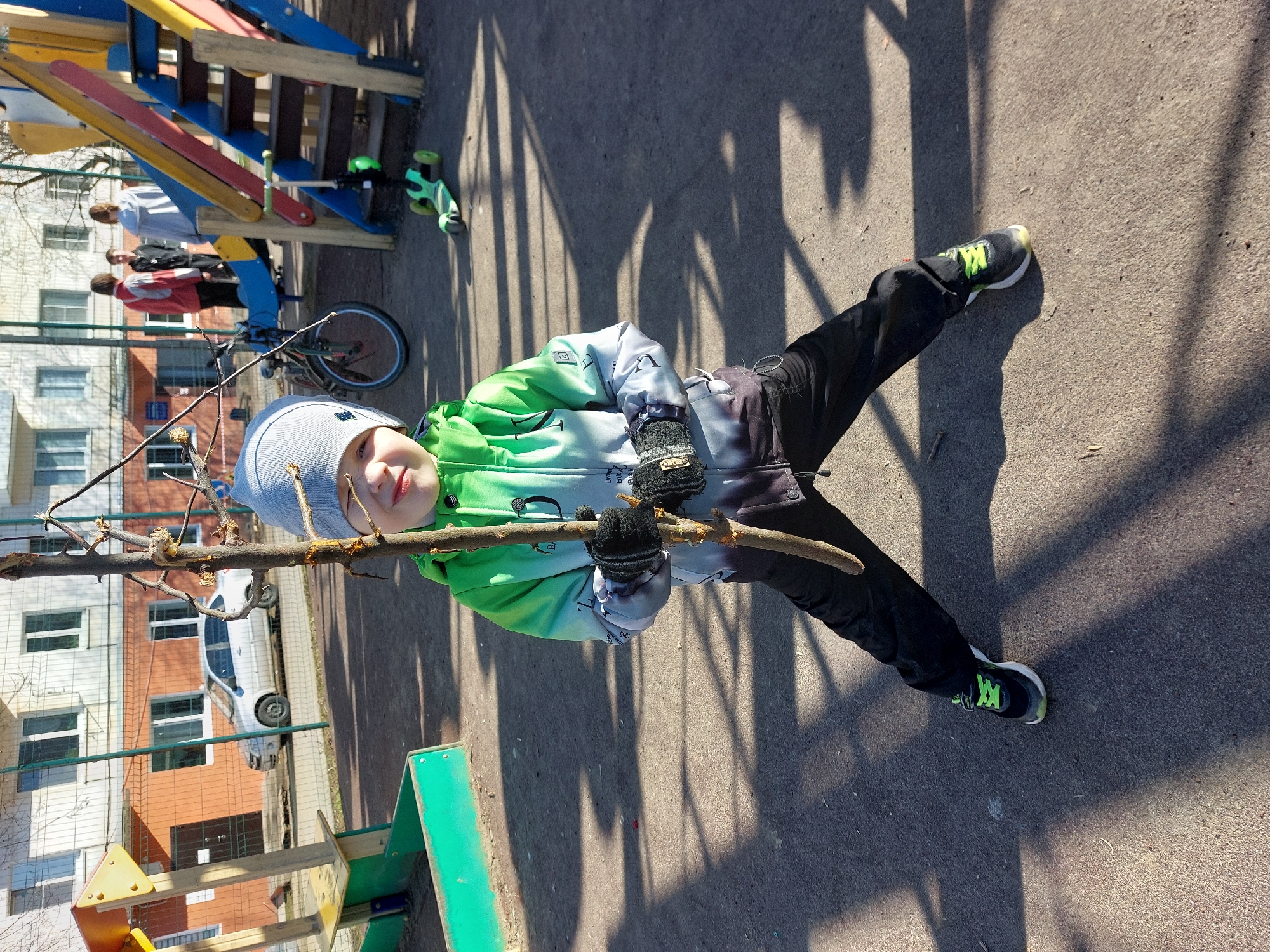 A young child, likely a boy, is the central subject of this brightly lit outdoor photograph, set in what appears to be a residential playground in Chaikovsky, Russia. He wears a light grey beanie, a green, grey, and black gradient jacket with stylized text, black gloves, black pants, and black sneakers with neon green laces. The child holds a substantial tree branch horizontally with both hands, looking directly at the camera with a calm, neutral expression, his legs spread apart.
In the mid-ground, a colorful playground structure with blue, red, and yellow elements is visible. Near it, three other individuals (adults or older children) are standing, and a bicycle is parked. A green scooter lies on the asphalt ground to the right. Further in the background, multi-story residential buildings and a white car parked on a street are seen, indicating an urban or suburban environment.
The ground is asphalt or concrete, with strong, elongated shadows cast by unseen objects, signifying a sunny day, likely mid-morning or afternoon. The child's attire, including a beanie, jacket, and gloves, suggests cool weather conditions. Partially legible, stylized text, including words like "Dream" and "ENJOY," is visible on the child's jacket sleeves, and "BA" on the chest area.