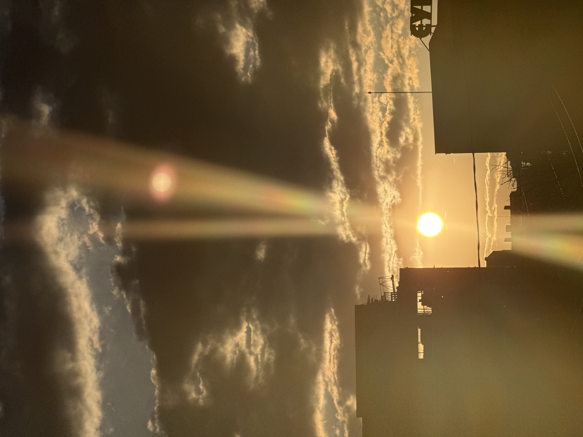 The image, oriented horizontally, depicts a dramatic sunset or sunrise over an urban or semi-urban landscape, identified by context as Vozdovac Urban Municipality, Serbia. The sky is a prominent feature, filled with a mix of dark and illuminated clouds. The sun, a bright white sphere, is positioned towards the right of the frame, low on the horizon, casting warm golden and orange light across the scene. Intense lens flares emanate from the sun, creating prominent light rays stretching horizontally and vertically across the sky.
Various buildings and structures are silhouetted against the bright horizon on the right and bottom edges of the image. These appear to be multi-story structures, possibly industrial or commercial, with visible roof elements and what might be antennae or construction frameworks. A slender streetlight or utility pole is also visible in silhouette against the radiant sky. In the upper right section, atop a building, there is a dark, partial silhouette of letters, possibly forming an unclear sign such as "CYH".
The scene captures the powerful interplay of natural light with cloud formations and urban architecture, characteristic of either dawn or dusk, creating a striking contrast between the bright sky and the darkened silhouettes of the man-made structures. The atmosphere is one of calm grandeur, with no human activity directly visible.