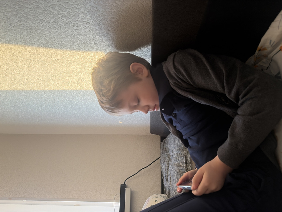 A young male child, estimated to be between 7 and 10 years old, with light brown hair, is depicted reclining on a dark gray upholstered sofa. He is wearing a dark blue collared polo shirt and a gray knitted cardigan, with dark trousers partially visible. The child holds a silver-framed smartphone horizontally with both hands, his gaze directed downwards at the device, indicating focused engagement. His facial expression is neutral with slightly narrowed eyes.
To the left of the child, a portion of a white window frame is visible. Below it, a black power strip or charging unit, with a black electrical cable plugged into it, is mounted against a light-colored, textured wall. A patterned pillow is partially visible on the sofa to the child's left. A prominent vertical band of golden sunlight illuminates a section of the textured wall to the right, suggesting direct light entry from an unseen window or light source. The environment is an indoor residential setting within Saint Petersburg, Russia.