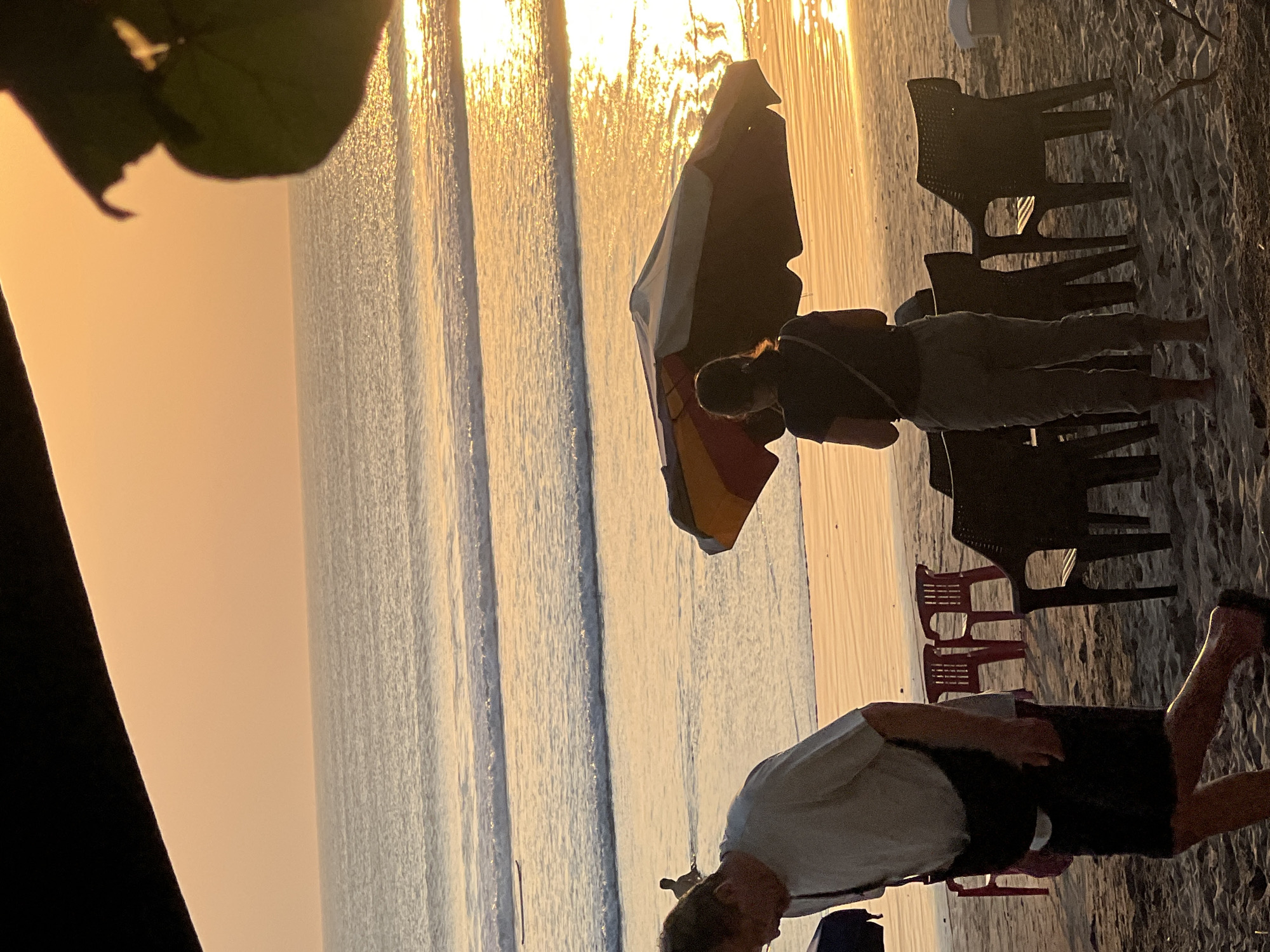 A beach scene unfolds at sunset in Kuta, Indonesia, with the golden light reflecting off the ocean and wet sand. In the foreground, a man in a light-colored t-shirt and dark shorts walks towards the left, his back mostly to the viewer, with a dark bag slung across his chest. To the right of the man, a woman stands facing the ocean, wearing a dark t-shirt and grey pants, with a dark bag over her shoulder. She is positioned near a colorful beach umbrella. Several plastic chairs, some red and some dark, are scattered on the sand, suggesting a seating area for beachgoers. The ocean exhibits gentle waves, and two figures are visible in the water further out, one appearing to be swimming and another on a surfboard. The sky is a gradient of orange and yellow, indicating the setting sun.