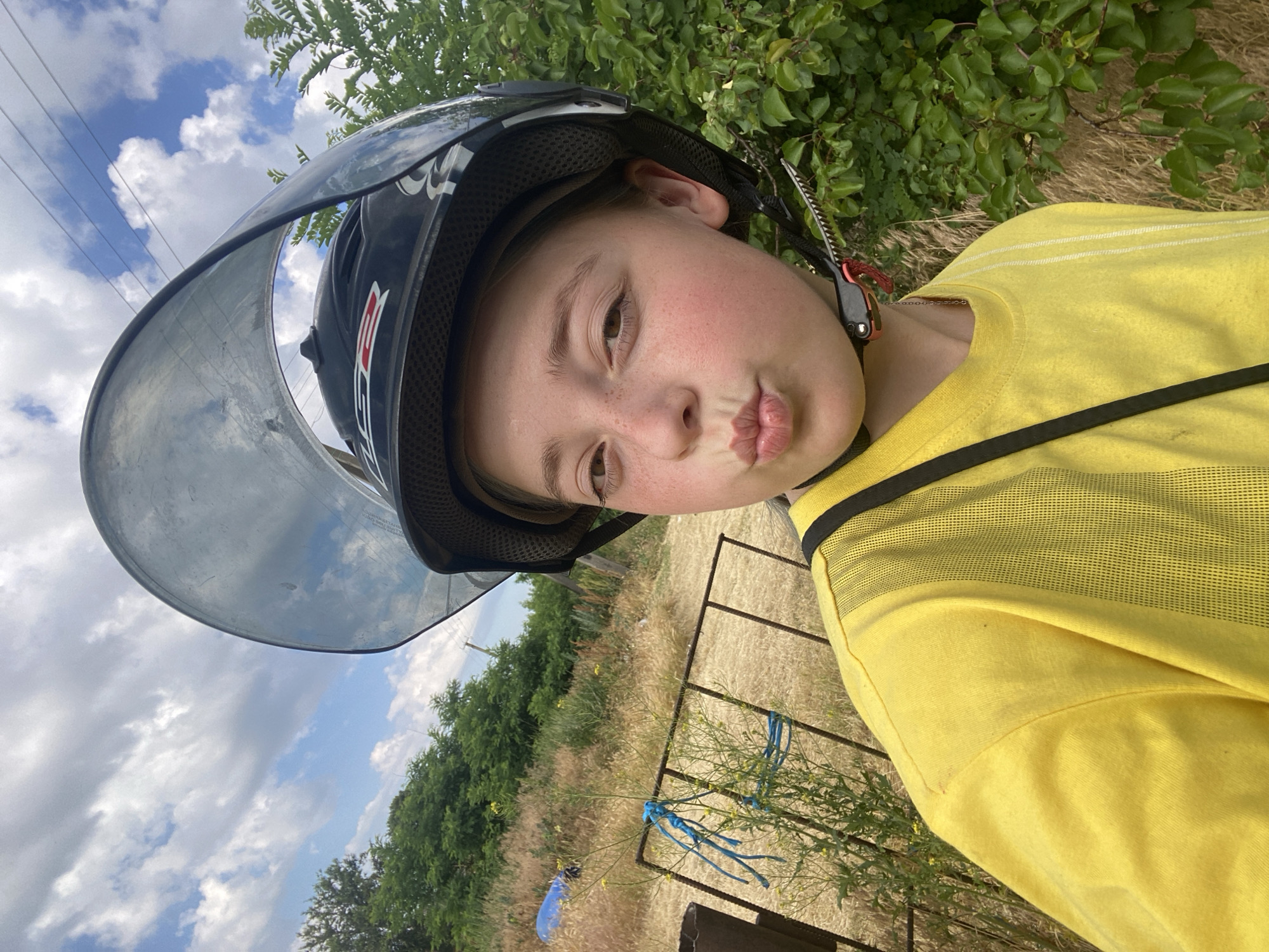 A young person, likely a child, is captured in a tilted, close-up selfie outdoors. They are wearing a black helmet with a clear visor and a bright yellow t-shirt. The person is making a puckered-lip expression, conveying a playful or pouty emotion. Their face is illuminated by natural light, suggesting it is daytime. Freckles are visible on their cheeks.
The background reveals a natural setting with greenery and dry grass, characteristic of a rural or outdoor environment. In the distance, trees and a fence are visible. The sky, with scattered clouds, is reflected in the helmet's visor. The image appears to have been taken during daylight hours, likely on a sunny day, given the bright lighting.
The setting is consistent with a location in Nova Praha, Ukraine, characterized by open fields and vegetation. There is no discernible text in the image. The overall impression is a candid, spontaneous moment captured by the young person themselves, showcasing their personality and surroundings.