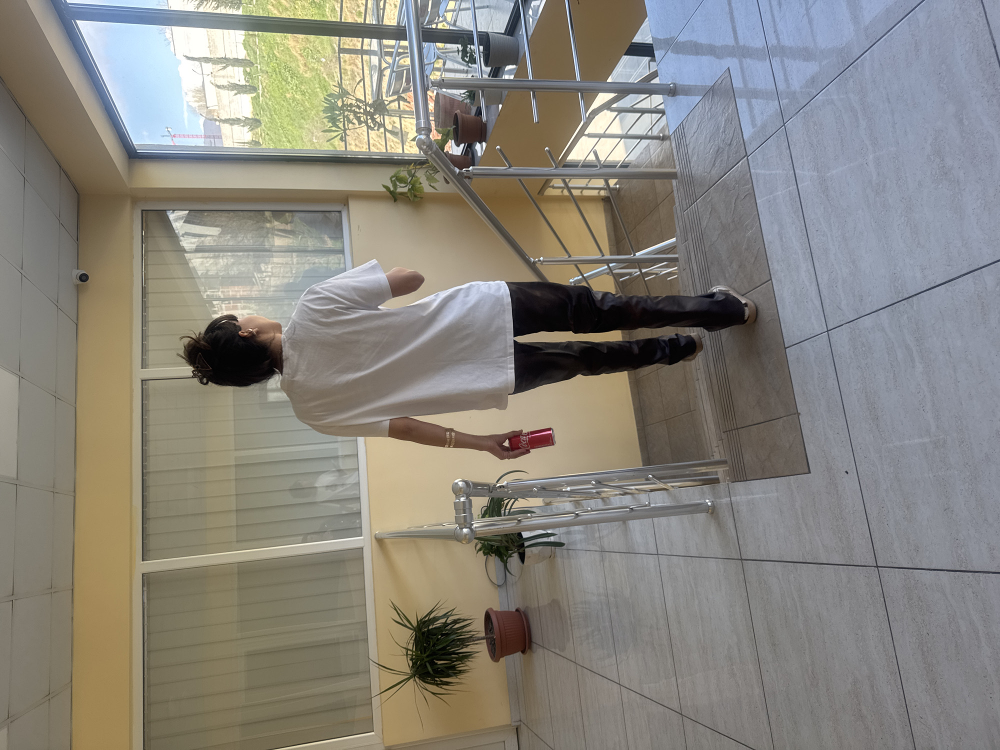 A medium shot shows a person standing inside a building near stairs. The person is wearing a white shirt and dark pants, holding a red Coca-Cola can in their right hand. The building appears to be an enclosed staircase or atrium with large windows and tiled floors. Plants in pots are positioned along the stairway and near the windows. Outside the windows, a view of Tbilisi, Georgia, is visible with trees and a building under construction.