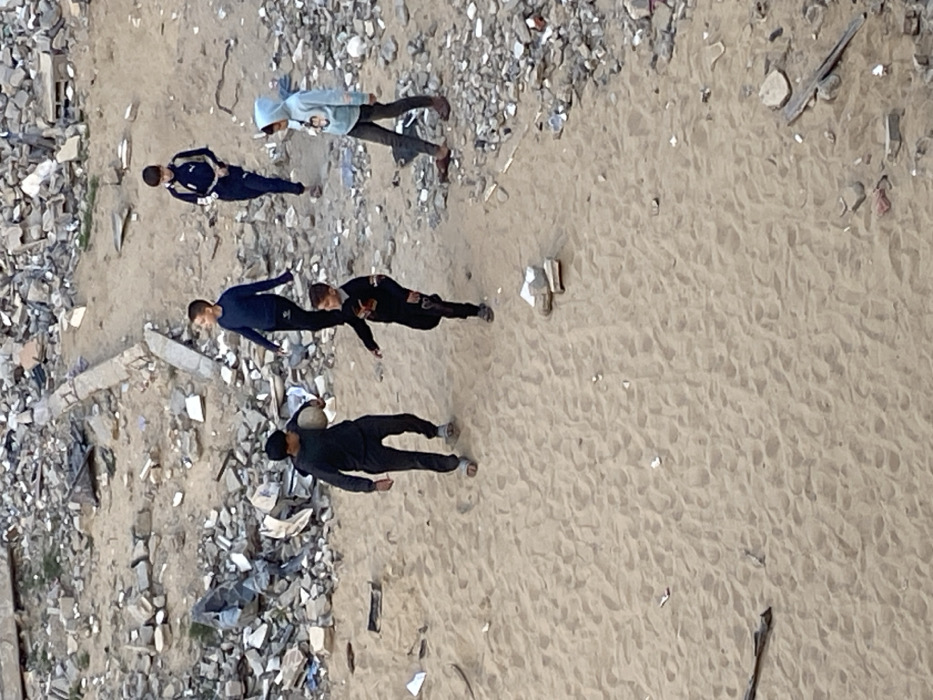 The image presents an aerial or high-angle view of five young individuals standing on a sandy area, which borders a vast expanse of debris and rubble. The scene suggests a desolate environment, possibly a demolition site, a neglected urban area, or a region affected by conflict.
The ground is sharply divided:
- On the left side, there is a large collection of broken concrete, rocks, and fragmented building materials, forming a barren and chaotic landscape.
- On the right side, a wide stretch of light brown sand covers the majority of the frame. Numerous footprints are visible on the sand, indicating recent activity. Scattered small pieces of litter, such as paper or plastic, can also be seen on the sandy surface.
Five individuals, appearing to be young males (children or teenagers), are present in the image. They are all standing on the sandy ground, generally positioned near the boundary with the rubble. Their attire is casual.
From left to right across the image:
1. The leftmost person wears a dark blue long-sleeved shirt and matching dark blue trousers. He stands facing slightly to the right of the frame.
2. Next to him, slightly further back, is another individual in a dark blue long-sleeved shirt, which features a white number "7" on the chest, and dark blue trousers. He also faces towards the right.
3. The third person from the left wears a dark black long-sleeved top and black pants. His head is turned towards the left side of the frame. His right arm is slightly extended downwards.
4. Standing directly to the right of this person is another individual, dressed entirely in black, including a long-sleeved top and pants. He is carrying a light-colored, possibly deflated, spherical object that resembles a ball, tucked under his right arm. He wears a dark cap or hat and light-colored sandals. He is facing towards the left.
5. The fifth individual, furthest to the right, is wearing a light blue hooded sweatshirt with the hood up, and dark pants. He also faces towards the left. He is wearing brown shoes.
The individuals do not appear to be engaged in a specific, unified activity. They seem to be standing, possibly conversing, or in the process of moving through the area. The presence of the ball might suggest recreational activities, though the overall backdrop is somber.
The lighting indicates it is daytime, with soft shadows suggesting either morning or afternoon. There are no clear indications of extreme weather conditions. No discernible text is visible in the image. The general impression is one of human presence in a harsh, possibly damaged, landscape.