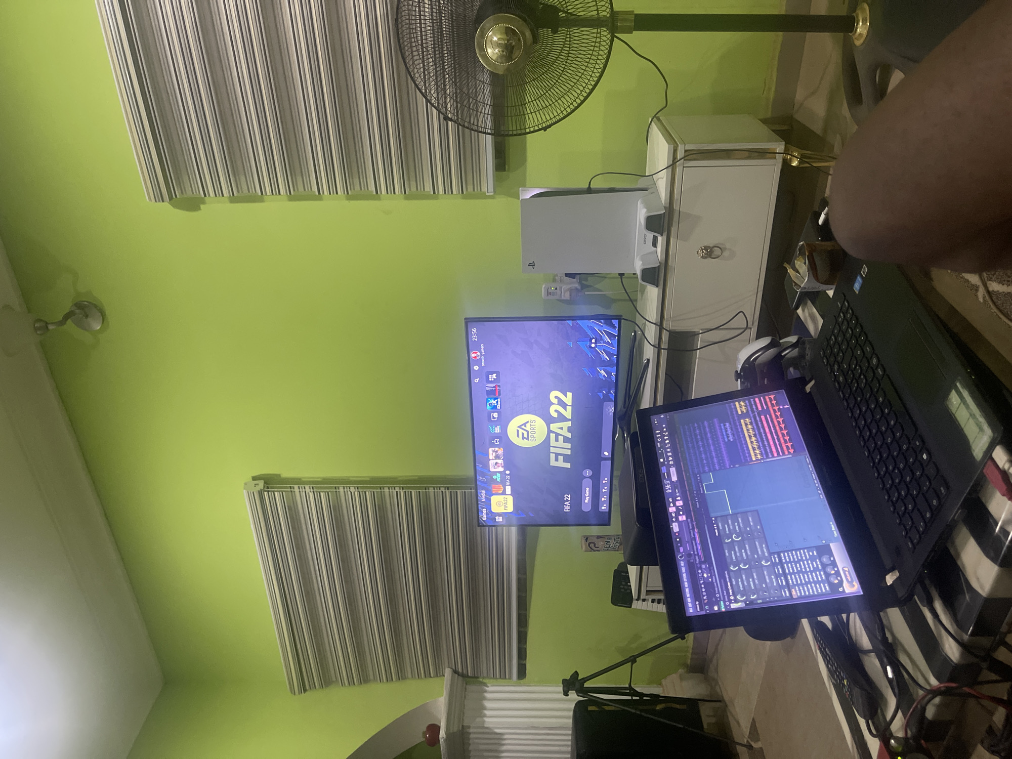 The image shows an indoor scene in a room with green walls. A standing fan is visible near a window covered with striped blinds. A TV displaying the game FIFA 22 sits on a white cabinet, alongside a PlayStation 5. A laptop, open and displaying a music production interface, sits on a striped surface. The lower part of a person's leg is visible on the right side of the frame. The OCR indicates text related to the FIFA 22 game on the TV screen and the time as 23:56. There's no information in the image that would indicate its location is specifically in Abuja, Nigeria.