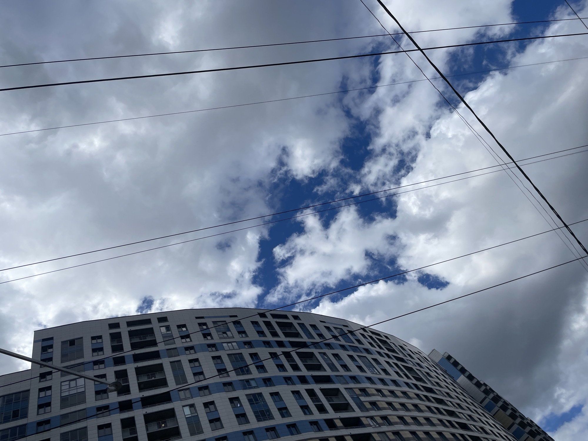 This is an upward-angle shot of a modern, multi-story building with a curved facade, situated in the city of Saint Petersburg, Russia. The building's exterior is a grid of windows and light-colored panels with blue accents. The sky above is filled with a mix of white and gray clouds, with patches of bright blue sky visible. Several black electrical wires crisscross the frame diagonally against the sky. No people are visible in the image. The overall impression is one of urban architecture against a dramatic sky.