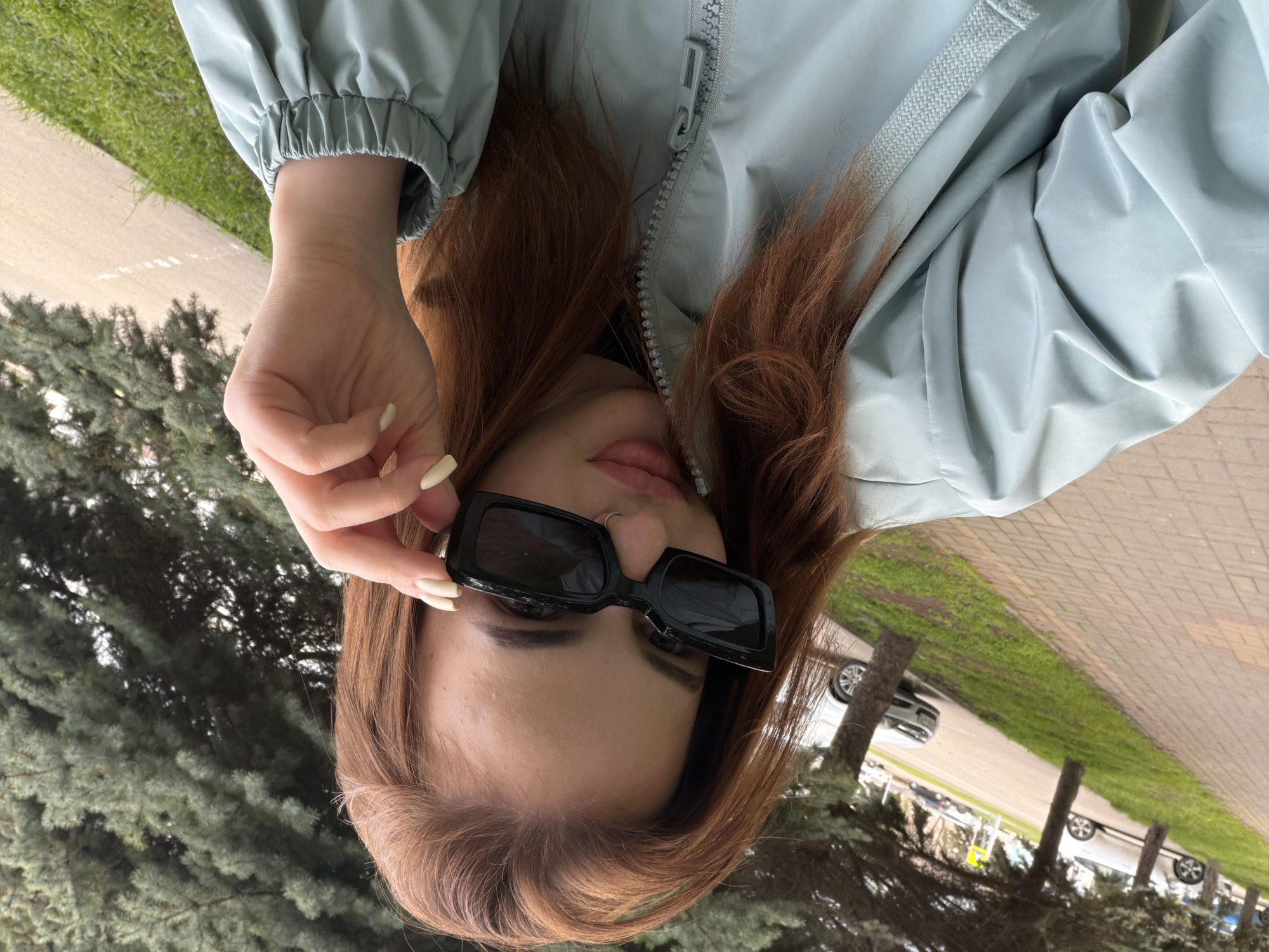 A young woman with long, auburn hair is the central figure. She is wearing a light blue jacket and square black sunglasses, which she is adjusting with her right hand. A nose ring is visible.
The background shows a sidewalk and grass, with trees and parked cars along the street. The scene appears to be in Novopokrovskaya, Russia.