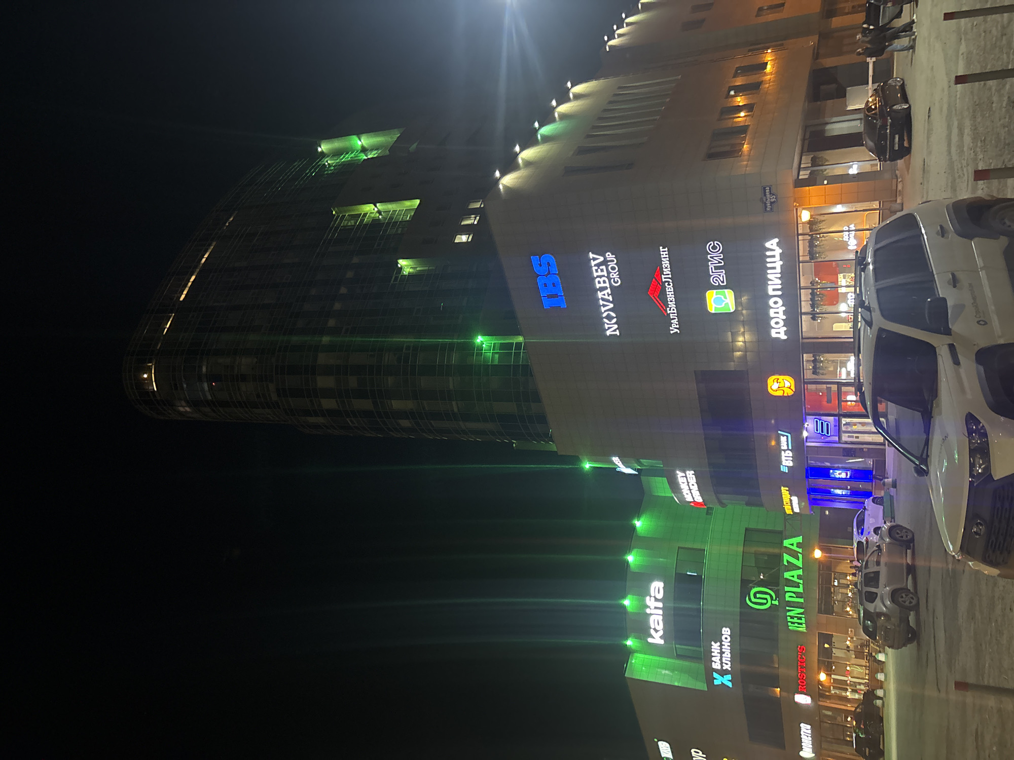 The image depicts a night scene of a commercial building in what appears to be a city environment, likely taken in Perm, Russia, based on visible signs. The building features several business names illuminated in bright, contrasting colors, including "IBS," "NOVABEV GROUP," "УРАЛБИЗНЕС ЛИЗИНГ" (Uralbusiness Leasing), "2ГИС" (2GIS), "ДОДО ПИЦЦА" (Dodo Pizza), "ROSTIC'S," "GREEN PLAZA," "kaifa," "БАНК Хлынов" (Bank Khlynov), "MONKEY GRINDER," "монетка," "ВТБ БАНК" (VTB Bank). Street address signage indicates the location as Куйбышева, 95 (Kuybysheva 95). The image includes several parked cars and at least one person is visible on the sidewalk. The lighting is a mix of neon signs and streetlights, creating a vibrant urban atmosphere.