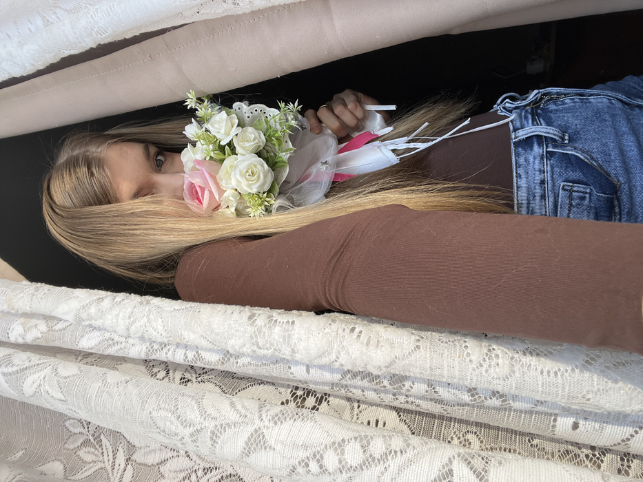 A blonde woman partially obscured by a floral arrangement of white and pink roses peeks out from beneath draped white lace fabric. She wears a brown, long-sleeved shirt and denim jeans. The scene is partially dark, creating a sense of enclosure. The woman's hair is partially covering her face, and she is looking directly at the viewer with one visible eye.