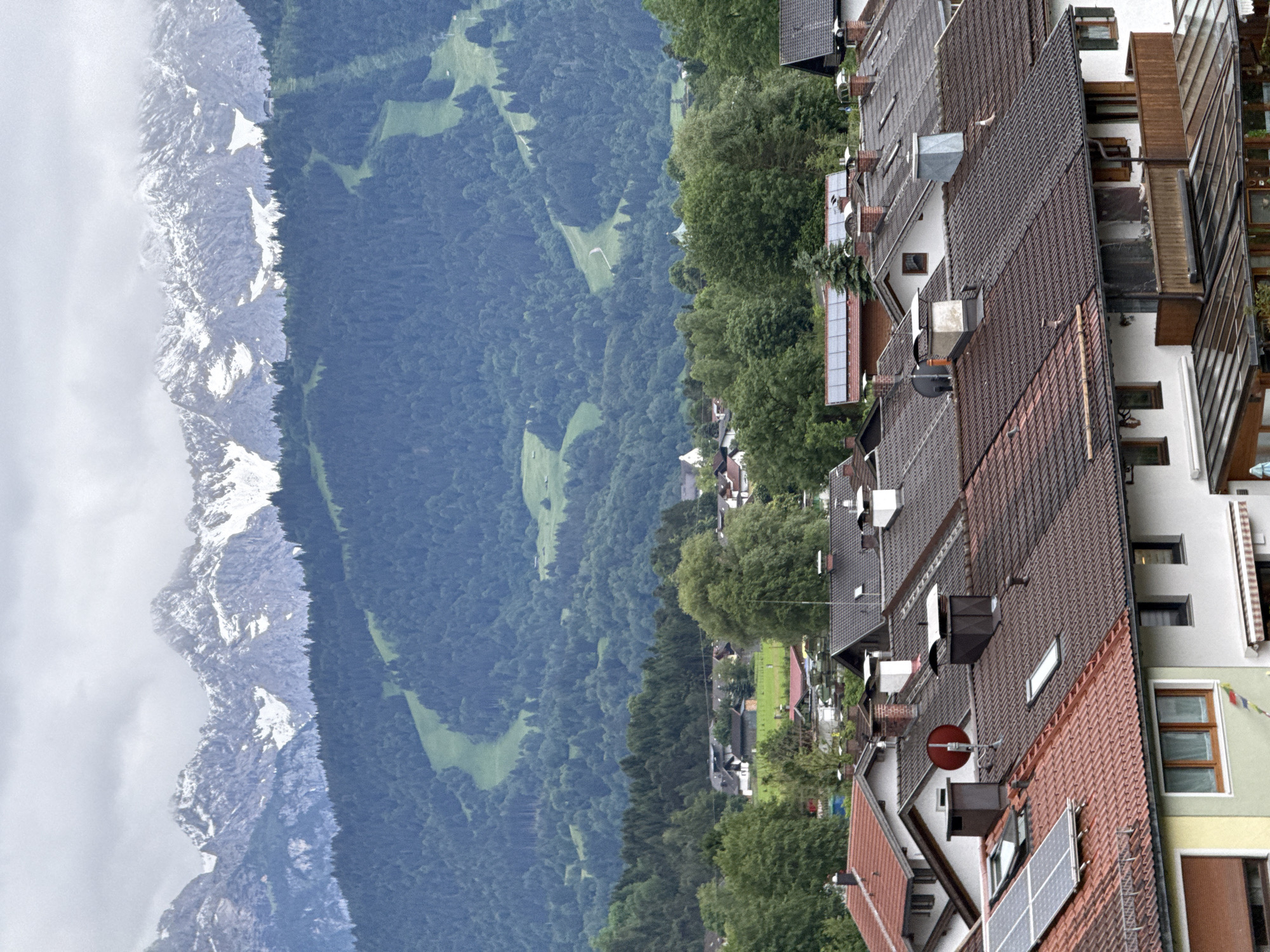 The image shows a panoramic view of Garmisch-Partenkirchen, Germany, with the Alps in the background. The foreground is dominated by the rooftops of residential buildings, many of which are covered in red or brown tiles. Some houses have solar panels installed on their roofs, while others feature prominent chimneys. A few buildings have balconies, and some have awnings. In the mid-ground, there are more houses nestled amongst lush green trees, suggesting a suburban or village setting. A small white church spire is visible above the trees. The background is dominated by a densely forested mountain slope, with a ski slope clearly visible as a lighter green path winding down the trees. The very top of the image shows snow-capped mountain peaks under a cloudy sky. The overall scene depicts a peaceful and picturesque Alpine town.