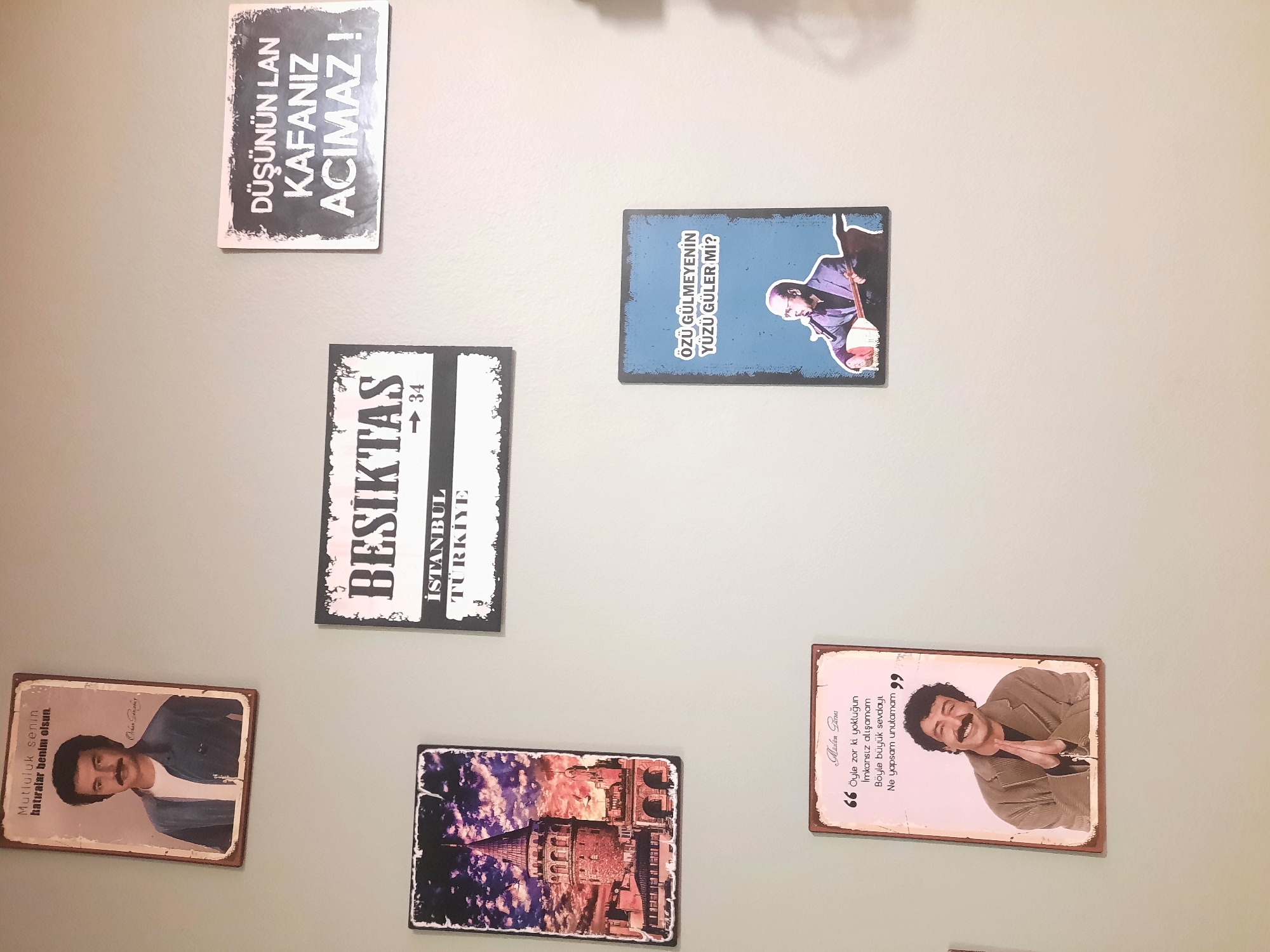 The image displays a wall adorned with several framed prints. At the top left, a framed print features a man with a mustache, dressed in a blue shirt and white t-shirt, with Turkish text above him. To its right, a rectangular print with a black border displays "BESIKTAS ISTANBUL TURKIYE" and the number "34". Further to the right, another framed print has the text "DÜŞÜNÜN LAN KAFANIZ ACIMAZ !" in white on a black background.
Below the "BESIKTAS" print, on the left, is a print depicting the Galata Tower in Istanbul against a dramatic sky. To the right of this, a vertical print with a blue background shows a man in a suit playing a stringed instrument, with the Turkish text "ÖZÜ GÜLMEYENİN YÜZÜ GÜLER Mİ?" above him.
At the bottom left, a framed print with a light pink background contains Turkish text and a portrait of a smiling man with a mustache, his hands clasped. The text appears to be a quote.
This scene is not located in Rosenheim, Germany. The prints themselves feature Turkish imagery and text, suggesting a Turkish cultural theme. The lighting indicates an indoor environment, likely a room with a plain, light-colored wall. There are no people present in the immediate scene other than those depicted in the prints.