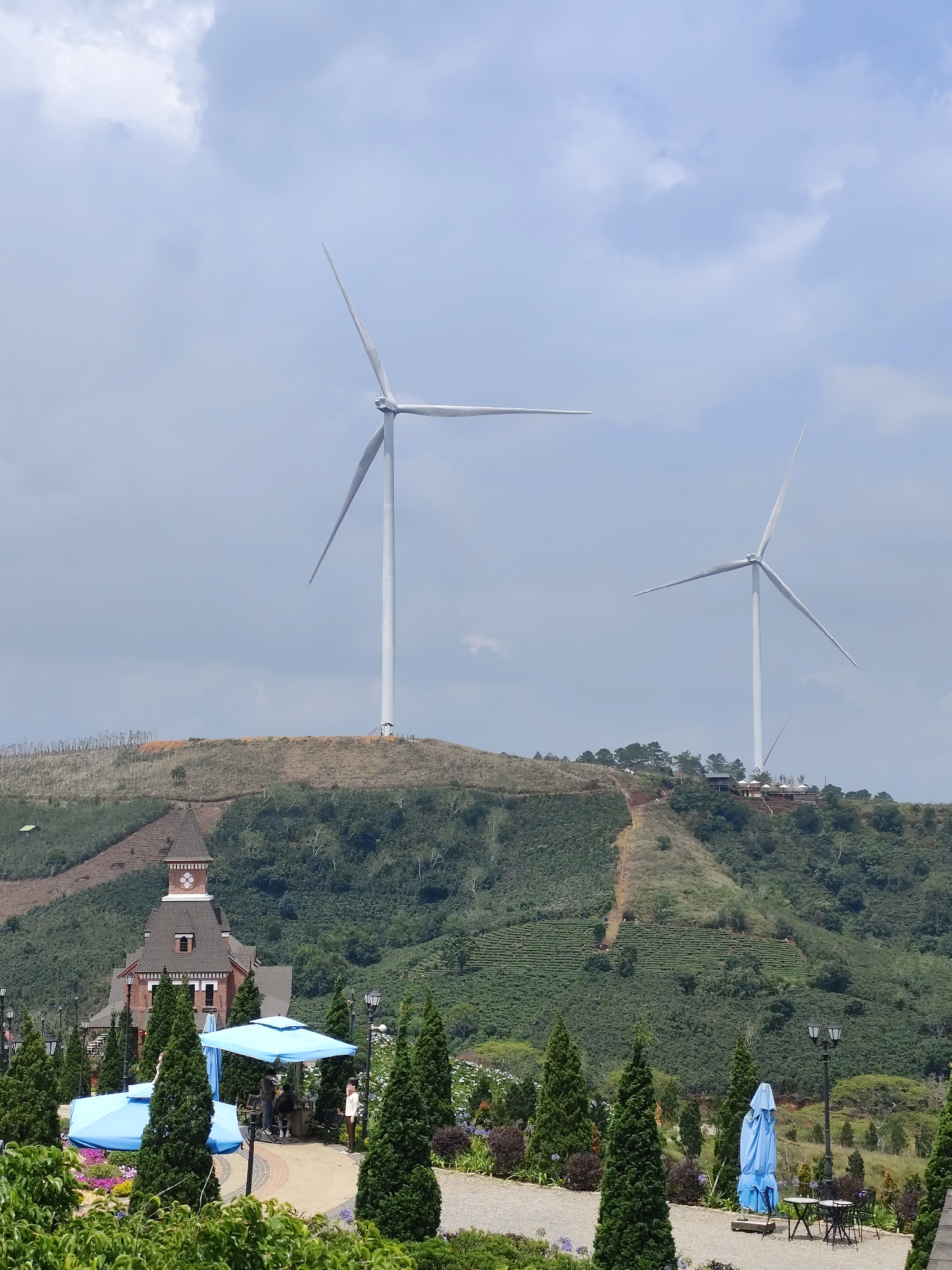 The image shows a landscape in Trường Thọ, Vietnam. Two large wind turbines stand atop two separate hills covered in greenery. The closest hill has a building with a clock tower and various blue umbrellas and trees in the foreground. A pathway can be seen leading up the hill to a cafe or restaurant. The sky is overcast.