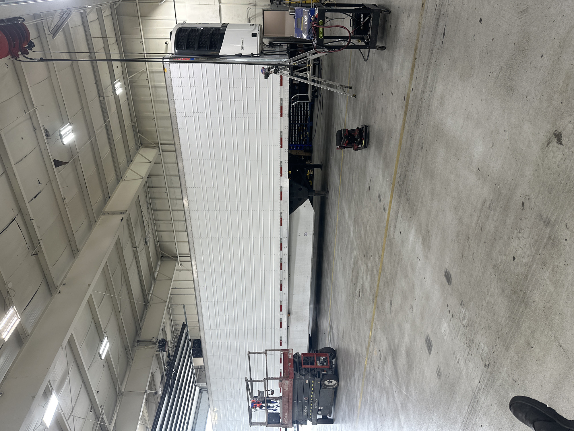 The image shows the interior of a large industrial warehouse, possibly a repair shop, located in Burr Ridge, Illinois, USA. A long, white refrigerated semi-trailer is parked in the center of the frame, occupying a significant portion of the space. To the left of the trailer, a red and black Skyjack scissor lift is positioned, appearing to be in use or ready for use. On the right side of the trailer, a silver A-frame ladder stands upright, and a black rolling cart with various electronic equipment, including a blue testing device, is situated nearby. There are no people clearly visible in the image. The warehouse has a high ceiling with exposed beams and fluorescent lighting. The floor is concrete, showing tire marks and some scuffing. The overall scene suggests maintenance or repair work being conducted on the semi-trailer.