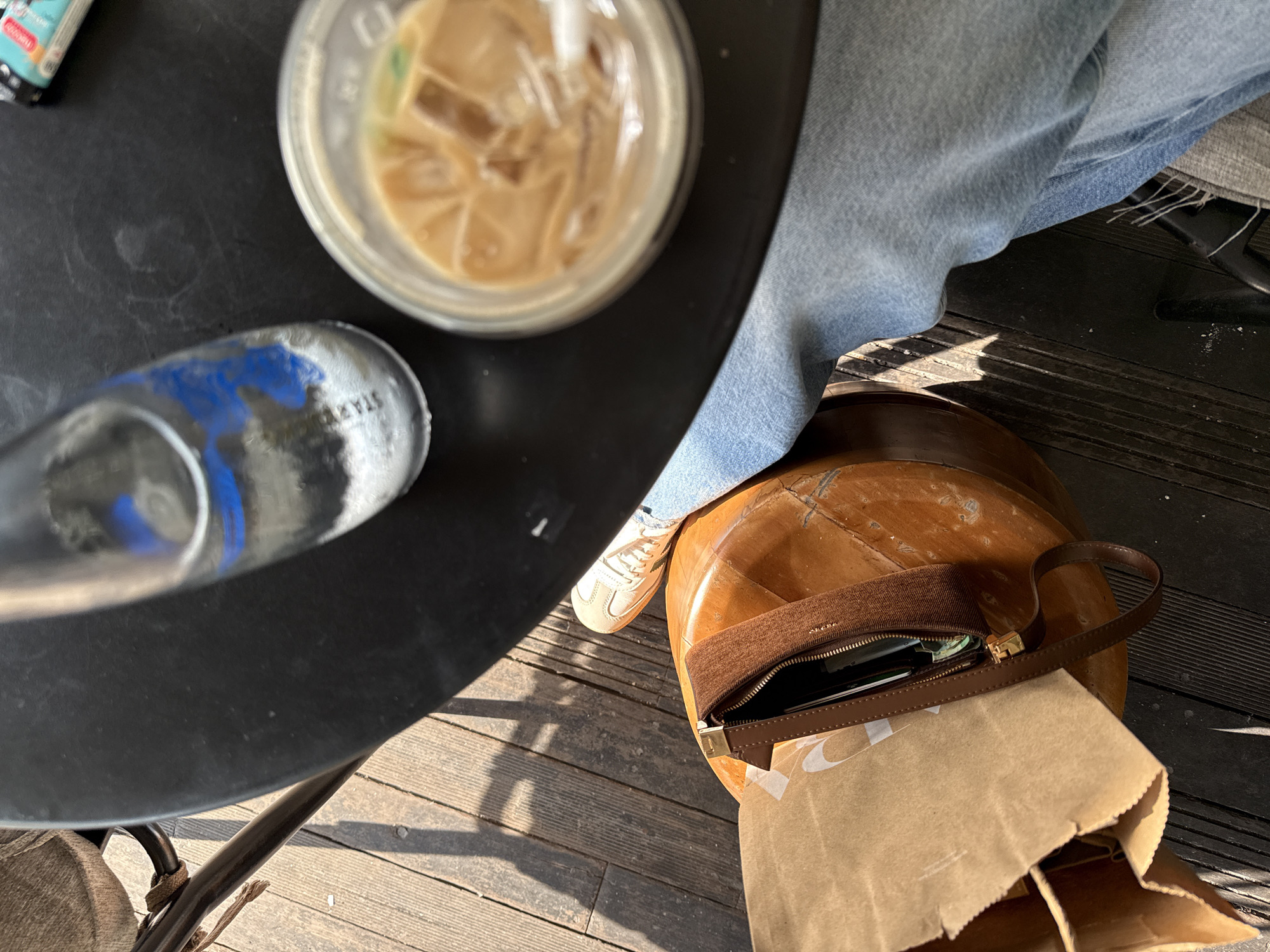 The image captures a first-person, overhead perspective of a casual outdoor setting in Kuta, Indonesia. The primary subjects include a person's legs and feet, a table with drinks, and a handbag.
A person is seated, with their legs visible, wearing light blue denim jeans and light-colored sneakers. They are positioned next to a round, black table. On the table, there is a clear plastic cup filled with an iced, light brown beverage, likely iced coffee, with a straw. Also on the table is a silver-colored water bottle with blue and white markings, possibly paint splatters or a design. The word "STAYL" is visible on the bottle.
Adjacent to the person and the table, on a wooden plank floor, rests a stylish brown handbag with a gold-colored clasp and a zipper closure. The bag appears to be made of a textured brown fabric and leather. Its main compartment is open, revealing contents like a mobile phone or wallet and what seems to be a white card. A brown paper bag with a partially visible white logo is positioned next to and partially underneath the handbag.
The scene suggests a relaxed outdoor break or meal. The strong directional light and distinct shadows cast by the table, person, and objects indicate bright sunshine, likely during the daytime, either morning or afternoon. The flooring consists of weathered dark wooden planks.