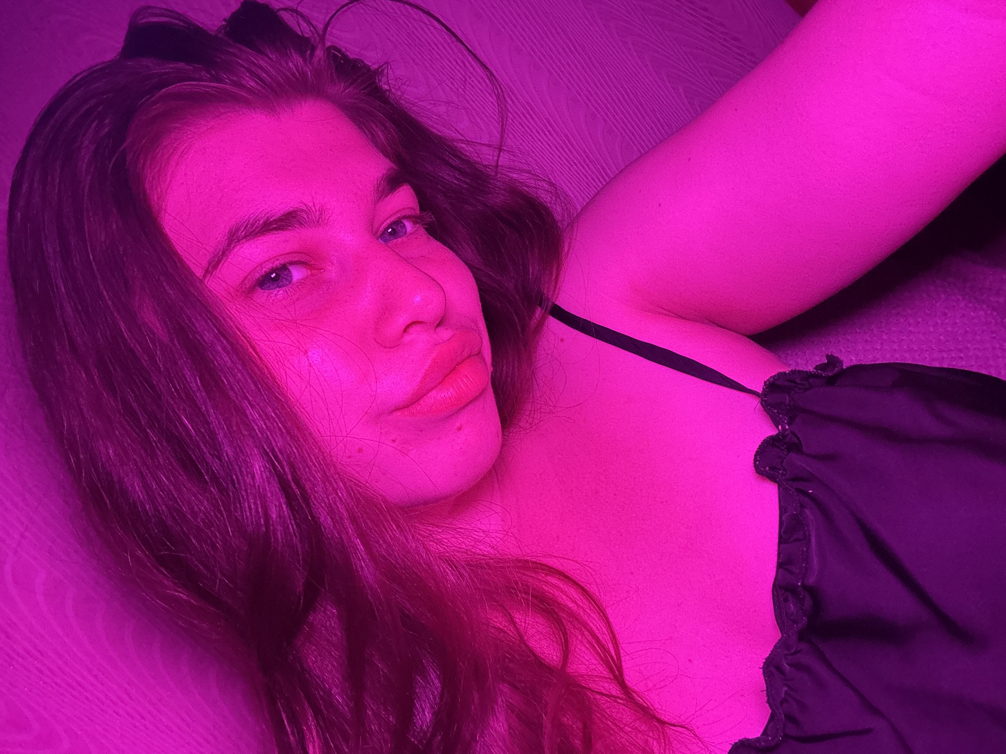 This is a medium shot selfie of a young Caucasian female with long, dark hair, wearing a black spaghetti strap top. She is looking at the camera with a soft expression, her head tilted slightly. The lighting is predominantly pink, casting a strong magenta hue over the entire scene. She appears to be indoors, with a textured surface visible behind her, possibly a wall or furniture. No specific details about the city of Мещёры, Russia are discernible from the image itself.