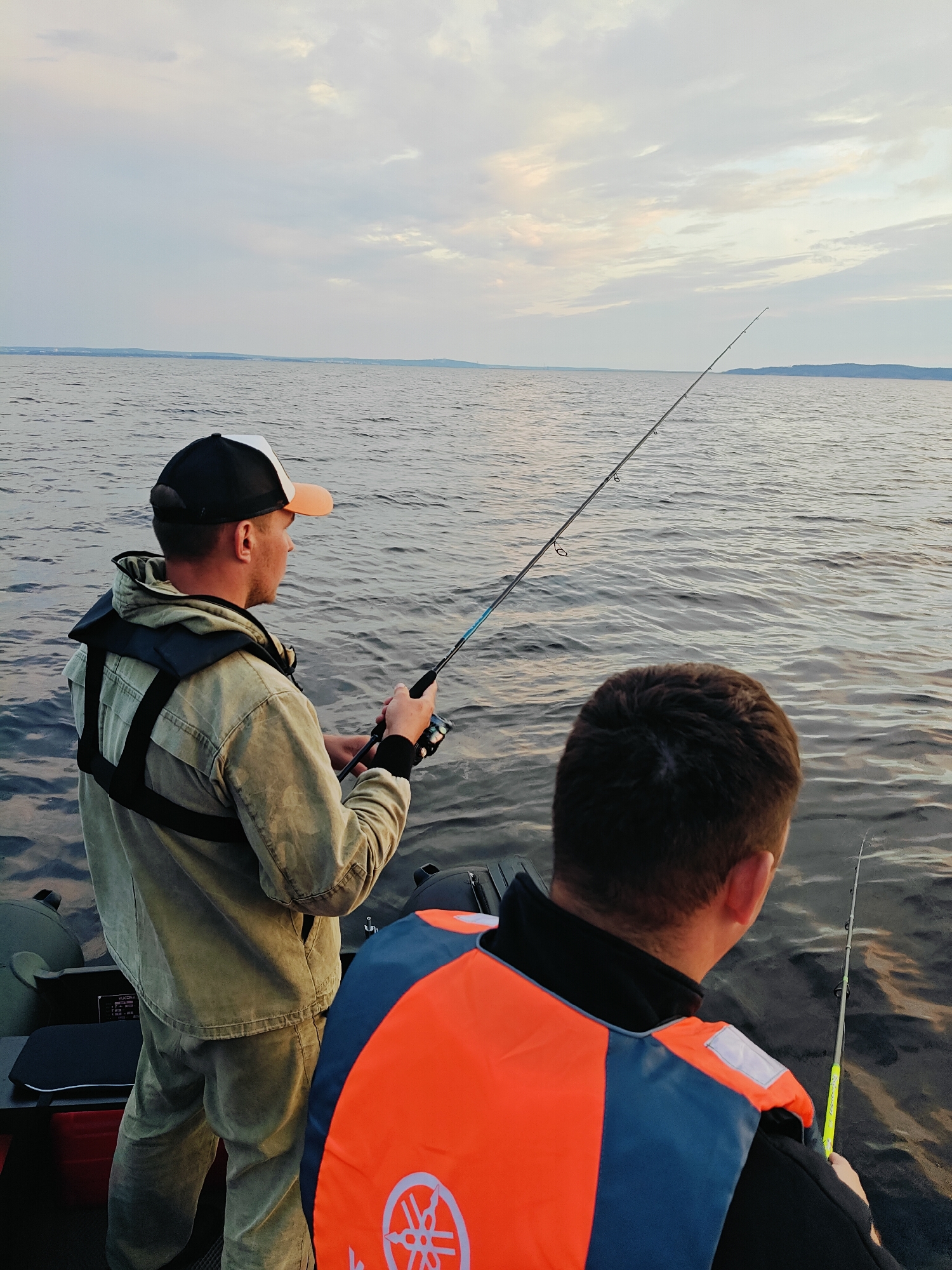 Two men are fishing from an inflatable boat on a large body of water. The man in the foreground is wearing an orange and blue life vest and is facing away from the camera, looking out at the water. The man in the background is wearing a light-colored jacket and a baseball cap, and he is holding a fishing rod. The water is calm with gentle ripples, and the sky is overcast with clouds. The location appears to be the Kama Reservoir, near the city of Chaykovsky, Russia.