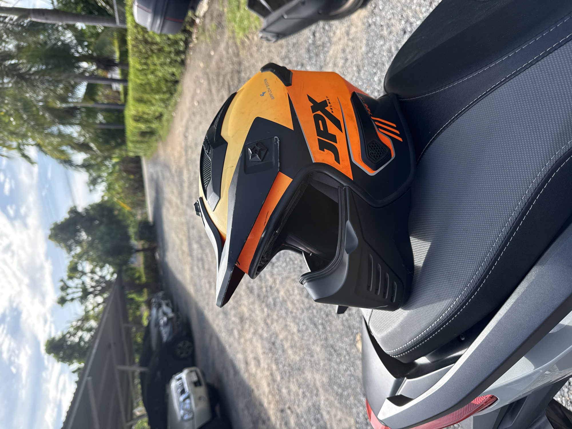 A motocross helmet, primarily orange and black with yellow accents and "JPX HELMET" branding, rests on the dark, textured seat of a motorcycle. The motorcycle is parked on a gravel lot, with a portion of its rear light and taillight visible in the lower left corner of the frame. In the background, cars are parked under a covered structure. Beyond the covered parking, a dirt road is visible, flanked by lush green bushes and palm trees, indicative of a tropical environment. The sky above is partly cloudy. The overall scene suggests a warm, sunny day in Ko Samui District, Thailand, possibly at a residential or resort area given the presence of covered parking and manicured landscaping. There are no people visible in the image.