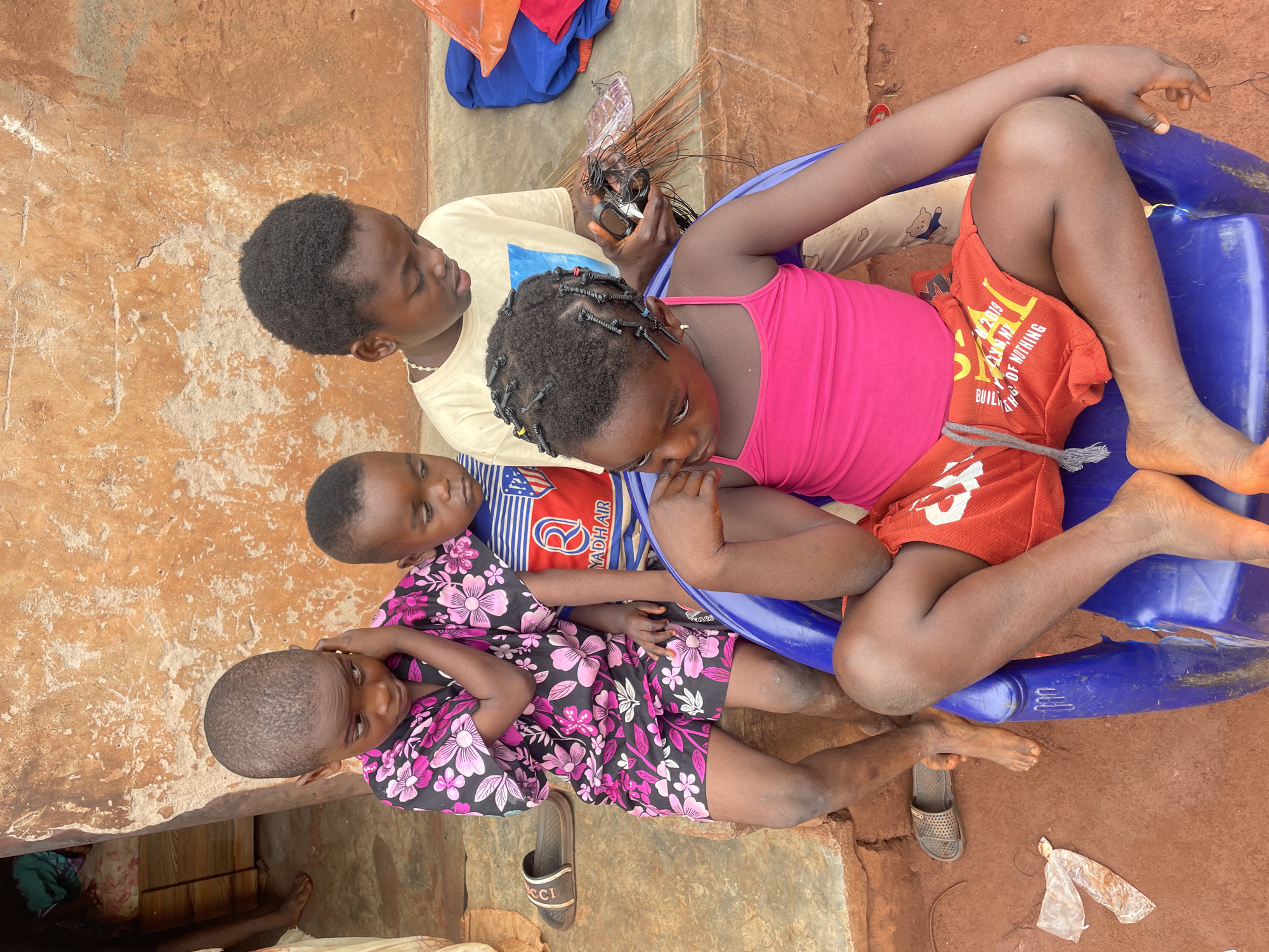 Four Black African children are seated together outside in Ogbe, Nigeria. One child wears a pink tank top and orange shorts, seated on a blue plastic object with their legs extended. A second child, wearing a tan shirt, sits beside them, holding a collection of wires and a small device. A third child, in a blue and red shirt, leans against the second. The fourth child, wearing a black floral pattern, is at the bottom of the group. A single sandal rests on the ground beside them. The ground is reddish-brown soil, and part of a concrete or stone structure is visible. A pile of clothes is next to the child holding the wires.