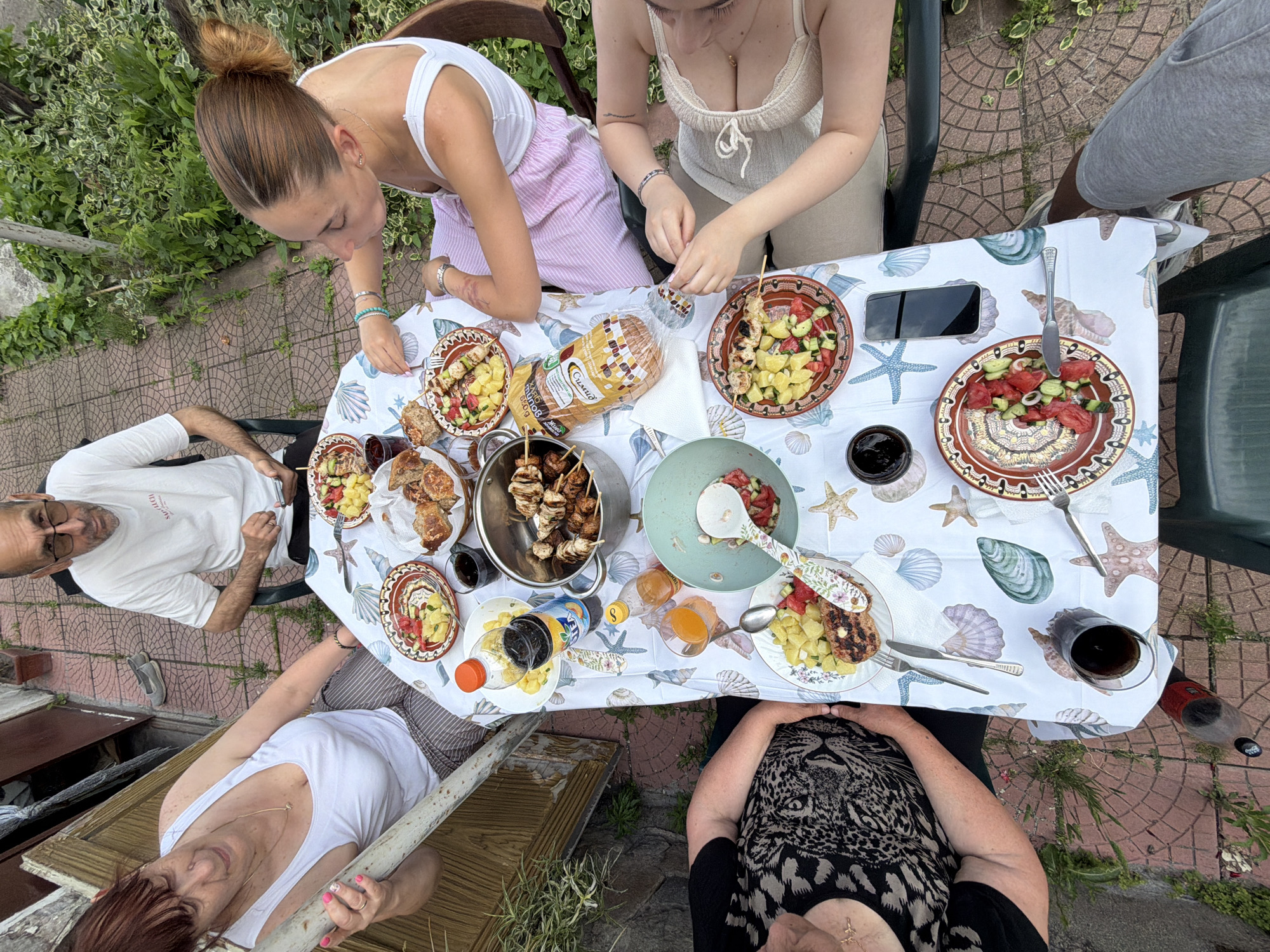 Five individuals are seated around a rectangular table outdoors on a patio in Krumovo, Bulgaria. The patio features reddish-brown interlocking paving stones and is bordered by green foliage. A weathered wooden structure is visible to the left.
The table is covered with a white tablecloth patterned with seashells and starfish. Numerous food items are arranged on the table, including:
- Multiple ceramic plates, some containing grilled meat skewers alongside a salad of diced potatoes and red vegetables (possibly tomatoes or watermelon).
- A large metal pot filled with various grilled meat skewers.
- A light blue bowl containing a salad of diced tomatoes and cucumbers, with a decorative serving spoon.
- A loaf of packaged Criso brand white bread.
- A plastic bottle of Fanta orange soda and another bottle containing yellowish oil or dressing.
- Several glasses filled with a dark liquid (likely cola or red wine).
- Assorted cutlery (forks, knives, spoons).
- A black smartphone positioned near the bottom right.
From left to right (clockwise):
1. An older woman with reddish-brown hair, wearing a white tank top and striped pants, rests her left arm on a light-colored wooden support. She wears a ring on her left ring finger and has painted pink fingernails.
2. A middle-aged man with short dark hair and glasses, wearing a white t-shirt, looks down at a smartphone he holds in his hands.
3. A younger woman with light brown hair tied in a bun, wearing a white tank top and light pink striped pants, looks down at the table. She has a tattoo on her right forearm and multiple bracelets.
4. A younger woman with light brown hair, wearing a light beige halter-neck top, uses her hands to engage with food on a skewer. She has a tattoo on her left forearm and multiple bracelets.
5. An older woman with dark hair, wearing a black and white leopard print top, is partially visible from the torso up.
A pair of grey slip-on shoes are visible on the paving stones behind the man. The scene depicts a casual outdoor meal.