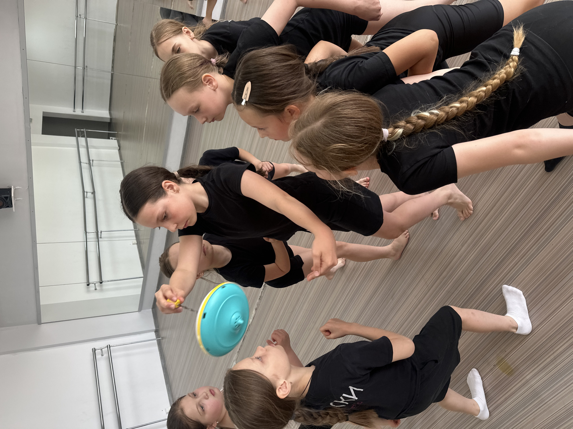 A group of young girls in black leotards are arranged in a line in a dance studio. They are mostly barefoot, except for one girl in white socks. The girls are looking down at a blue and yellow spinning top, which one of the girls is holding. The studio has mirrors and barres, which appear standard in dance studios. The location cannot be verified as Votkinsk, Russia.