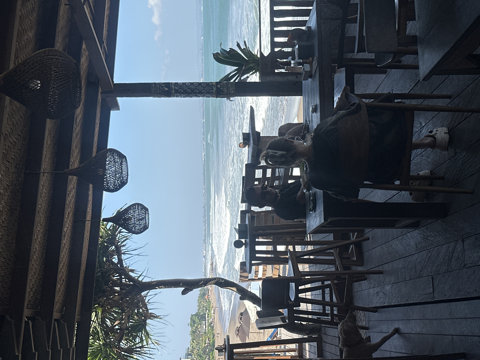 In North Kuta, Indonesia, a beachside open-air restaurant features wooden decking and a thatched roof. The foreground shows a dog walking on the wooden floor. Several wooden tables and chairs are arranged for dining. In the midground, two patrons are seated at a table, facing away from the camera, with a view of the ocean. A man with sunglasses is seated at another table. The background displays a sandy beach with umbrellas and people, leading to the turquoise ocean with rolling waves. Palm trees and lush vegetation are visible on the coastline, under a clear blue sky. Woven pendant lights hang from the ceiling.