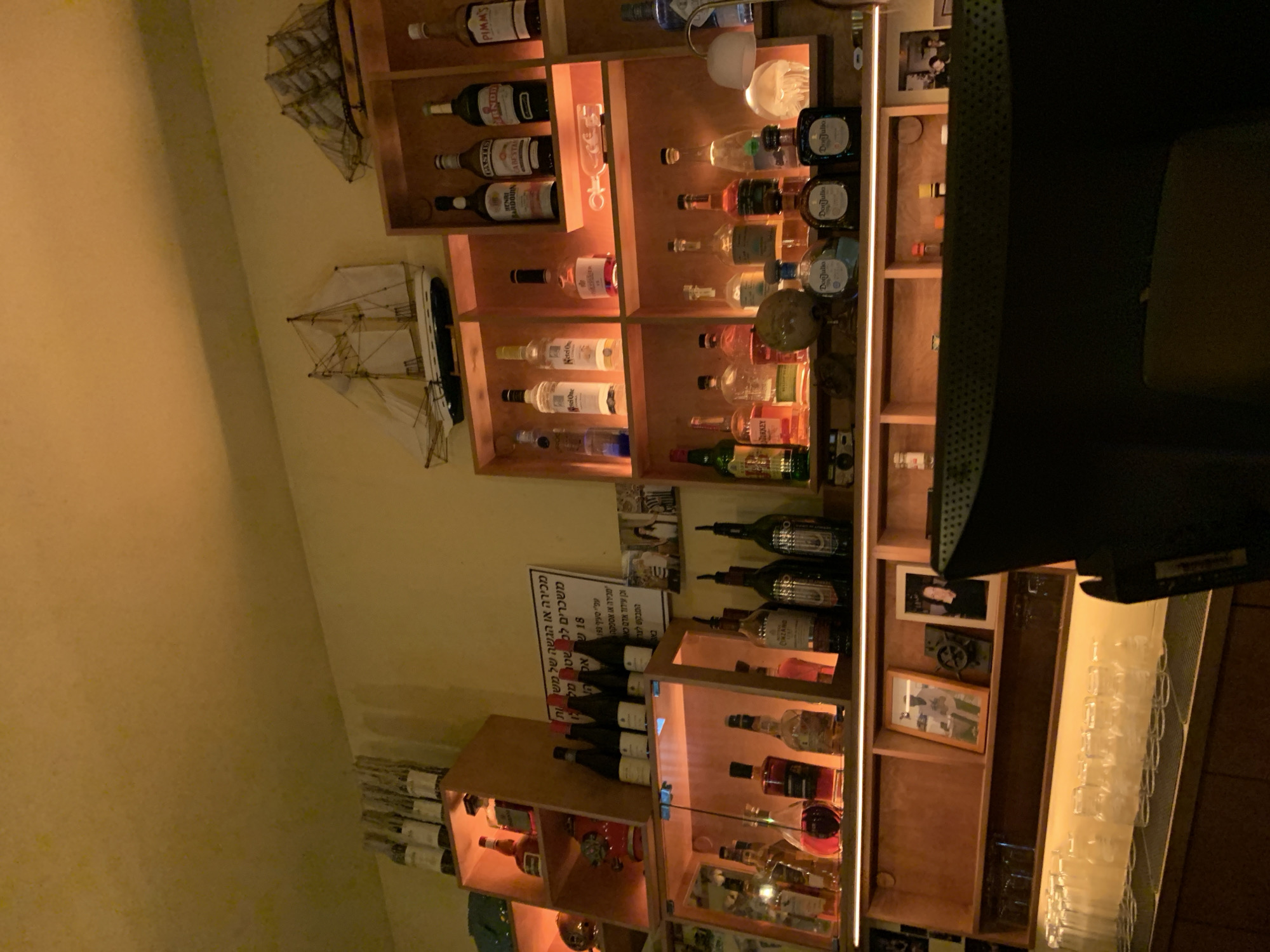 The image shows a bar interior in Tel-Aviv, Israel. It features shelves stocked with various bottles of alcohol, including brands like Pimm's, Don Julio, Ketel One, and Henri Bardouin Pastis. There are also wine bottles and model ships displayed on the shelves. A sign with Hebrew text is visible, referencing laws related to the sale or serving of alcoholic beverages to individuals under the age of 18. The bar is well-lit, with glasses hanging above a counter.