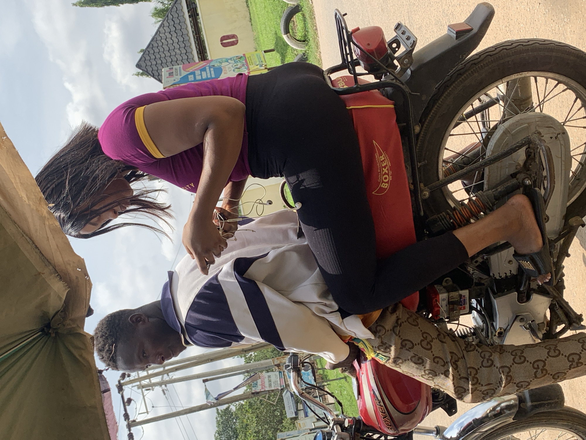 This media file captures a candid moment of two people on a red motorcycle, seemingly in Abuja, Nigeria. A woman, wearing a purple top with a yellow trim and black leggings, is leaning forward, her long dark hair obscuring her face. She is seated behind a man who is driving the motorcycle. The man is wearing glasses, a white and blue striped jacket, and patterned trousers that appear to be a luxury brand.
The motorcycle itself is red and has a prominent "BOXER" logo on a red bag attached to the rear. There are various other details visible, including parts of the engine and wheel. In the background, there's a glimpse of a building with a dark, pitched roof and some green foliage. A few signs with text are also visible, one appearing to advertise an "ACADEMY" and another mentioning "Orphan" with a phone number.
The image appears to have been taken during the daytime, with bright lighting and a cloudy sky suggesting it could be either morning or afternoon. The overall atmosphere seems casual and everyday, with no overt indication of a specific event. The orientation of the image suggests it was taken in a vertical format.