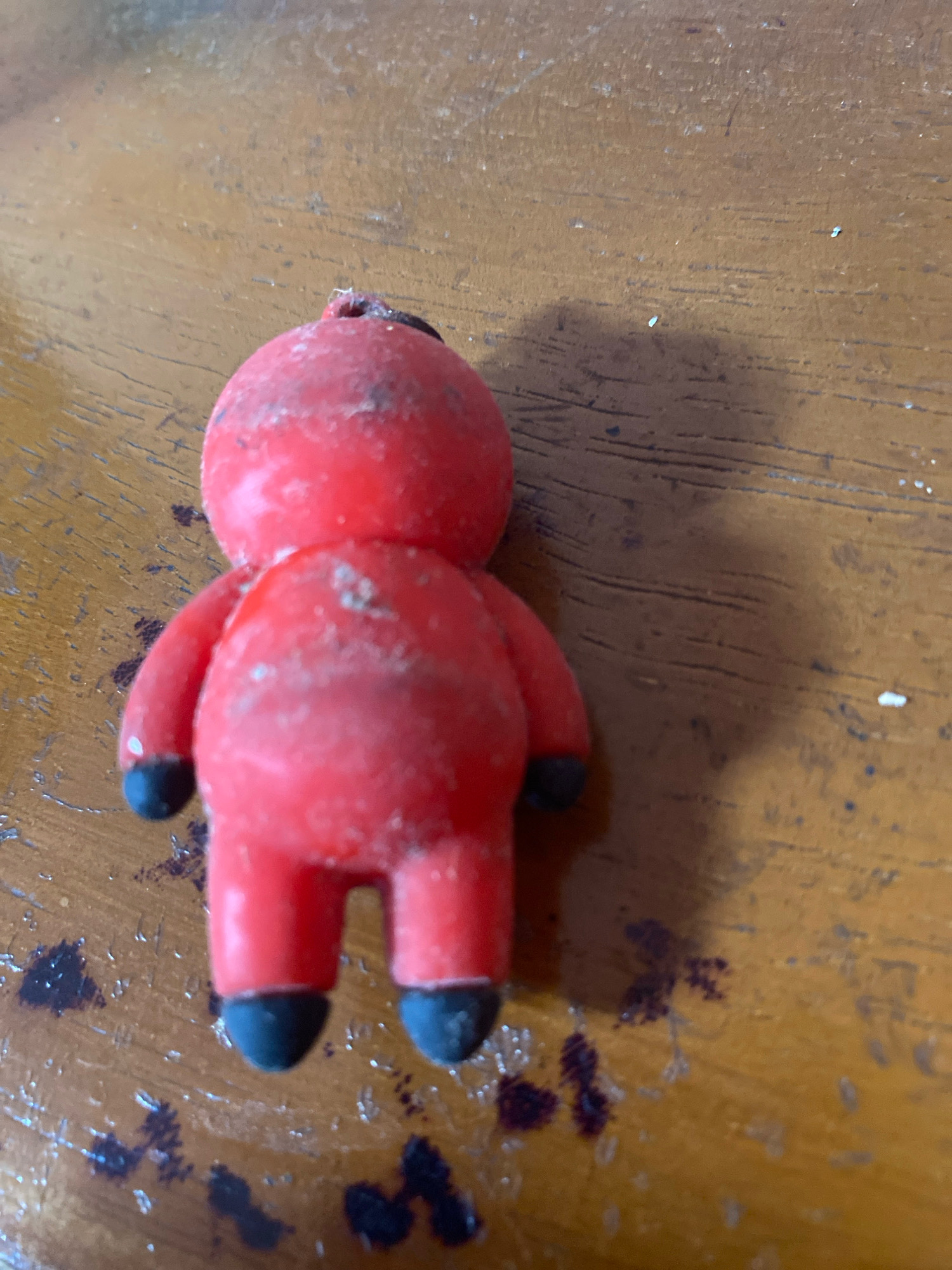 A small, red, anthropomorphic figure with black feet is positioned on a wooden surface. The figure appears to be made of a soft material, possibly wax or rubber, and has a rough, speckled texture. It has stubby limbs and a rounded head. The wooden surface is worn and has dark stains and lighter speckles scattered across it. The image is a close-up shot, with the figure occupying the majority of the frame. There is a shadow cast on the surface to the right of the figure. The scene is not identifiable as being in Mojo Pahit, Indonesia.