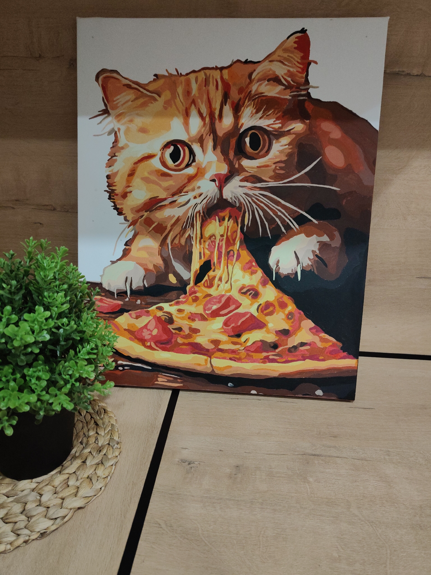 The image shows a painting of an orange cat eating a slice of pizza. The painting is on a canvas and is placed on a wooden surface. A small potted green plant is next to the painting. The painting depicts the cat with its eyes wide open and its paws resting on the pizza slice, with cheese dripping from its mouth. The pizza has toppings such as pepperoni and other unidentified ingredients. The background of the painting is a plain white color. The wooden surface appears to be a tabletop or a shelf. The location is Unknown, Unknown.