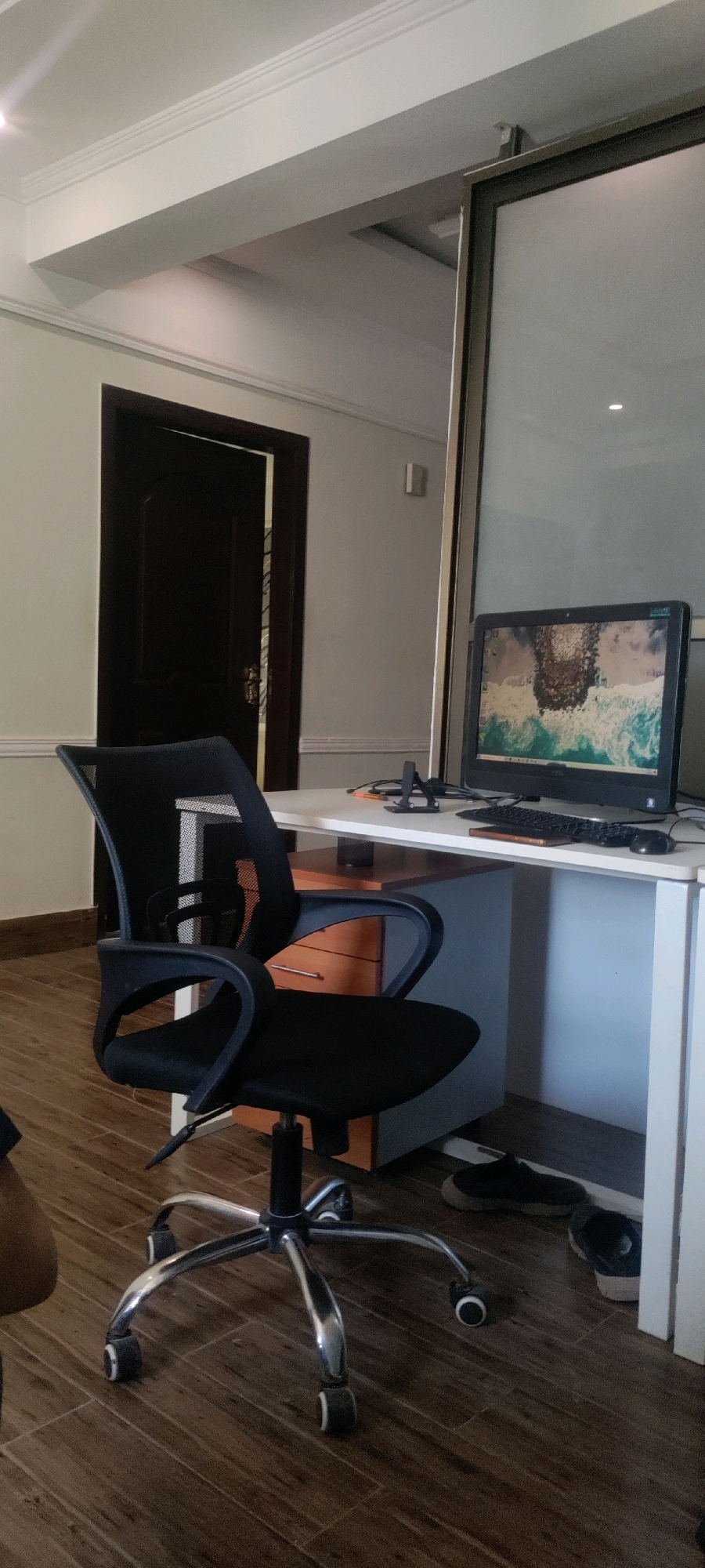 The image shows an indoor office setting with a dark wooden door slightly ajar on the left. In the foreground, a black mesh office chair with a silver base and wheels is positioned. To the right of the chair is a white desk. On the desk, there is a computer monitor displaying a landscape image, a keyboard, a mouse, a smartphone stand, and a phone. Beneath the desk, there is a set of drawers with orange fronts. The floor is made of dark wood planks. A large glass panel is visible on the right side of the frame, reflecting light. The overall environment appears to be an office space in Khana Dak, Pakistan. There are no visible people in the frame.