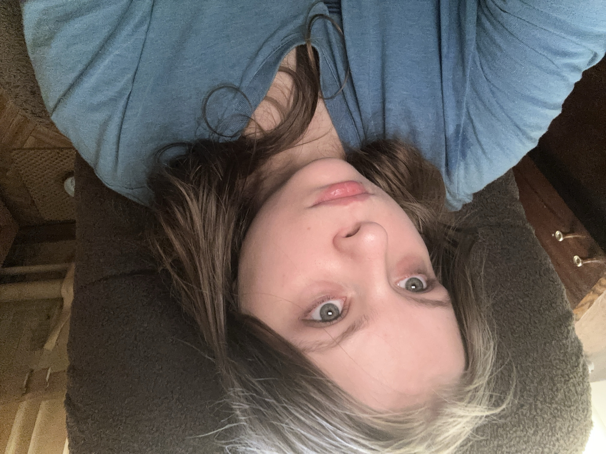 A young woman with fair skin, light green eyes, and brown hair with silver highlights is lying down. She is wearing a blue v-neck shirt. The surface she is lying on appears to be a brown upholstered cushion. A wooden object with drawers and knobs is visible in the background. The location in Moscow, Russia cannot be determined based solely on this image.