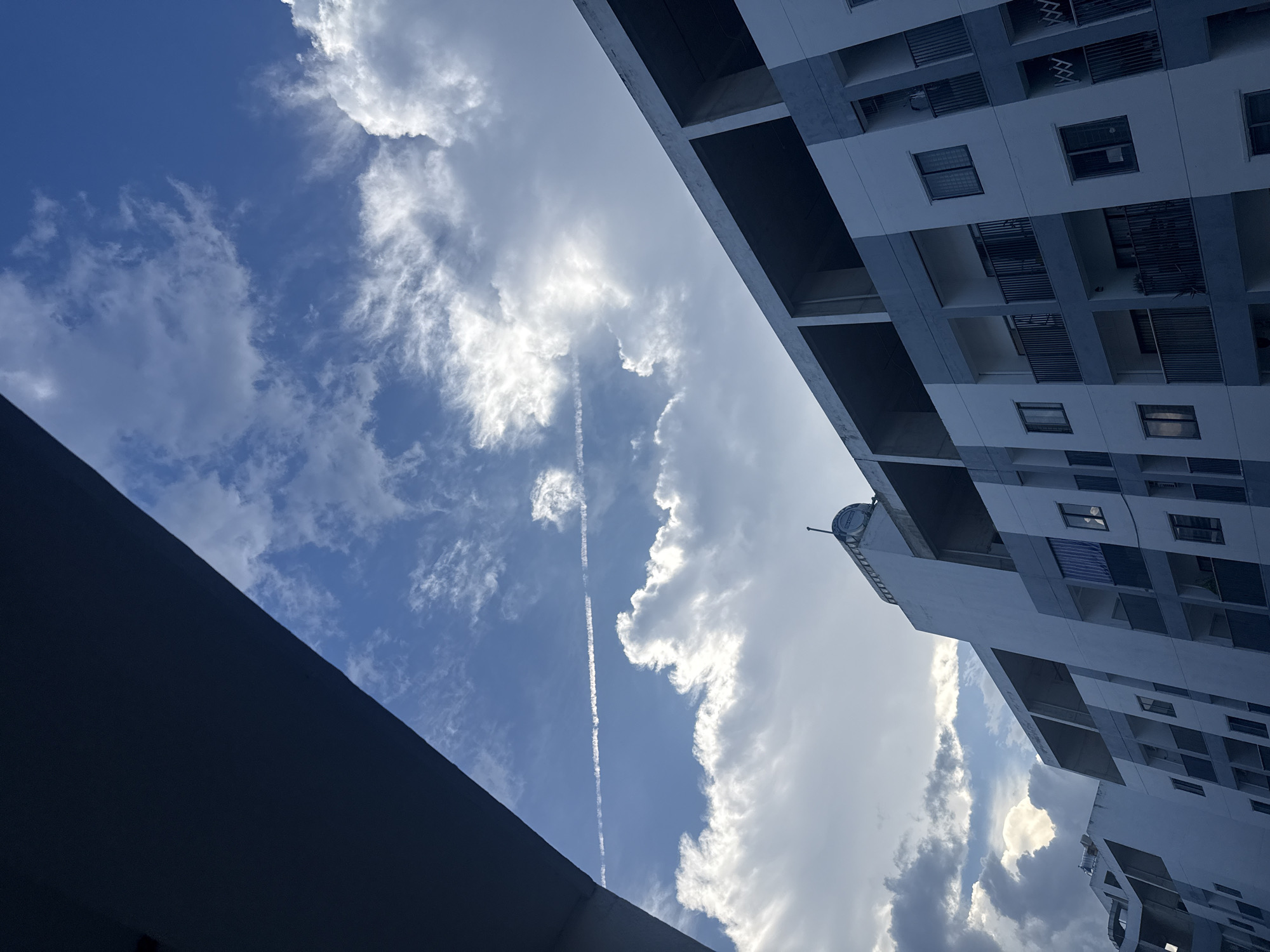 The photograph captures a view looking up at a modern apartment building against a dynamic sky. The building, with its distinctive grey and white facade and balconies, appears to be under construction or newly built, as some upper levels show exposed concrete. The sky is filled with a mix of white cumulus clouds and thinner, wispy clouds, with a distinct contrail from an aircraft streaking across the blue expanse. The perspective is from a low angle, with a dark, possibly overhang or architectural element, dominating the upper left corner of the frame. The lighting suggests it might be late afternoon or early evening, with soft light illuminating the clouds. Based on the architectural style of the building and the presence of lush vegetation visible in the background of some crops (though not clearly in the main image), this scene is likely situated in Nha Trang, Vietnam, a coastal city known for its contemporary developments and tropical climate. There are no people visible in the image.
